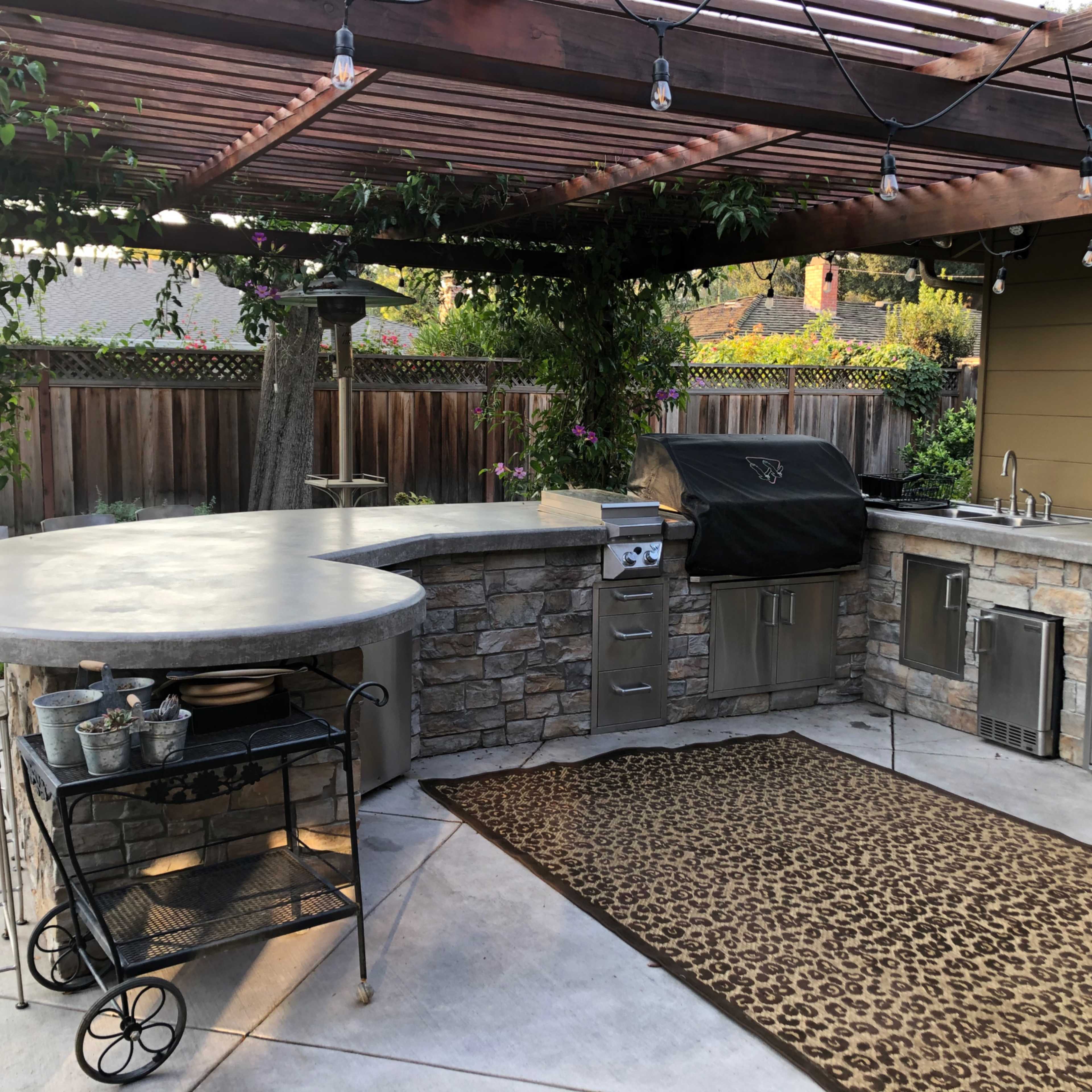 An outdoor kitchen area featuring a stone countertop, a grill, an outdoor sink, and seating under a wooden pergola.