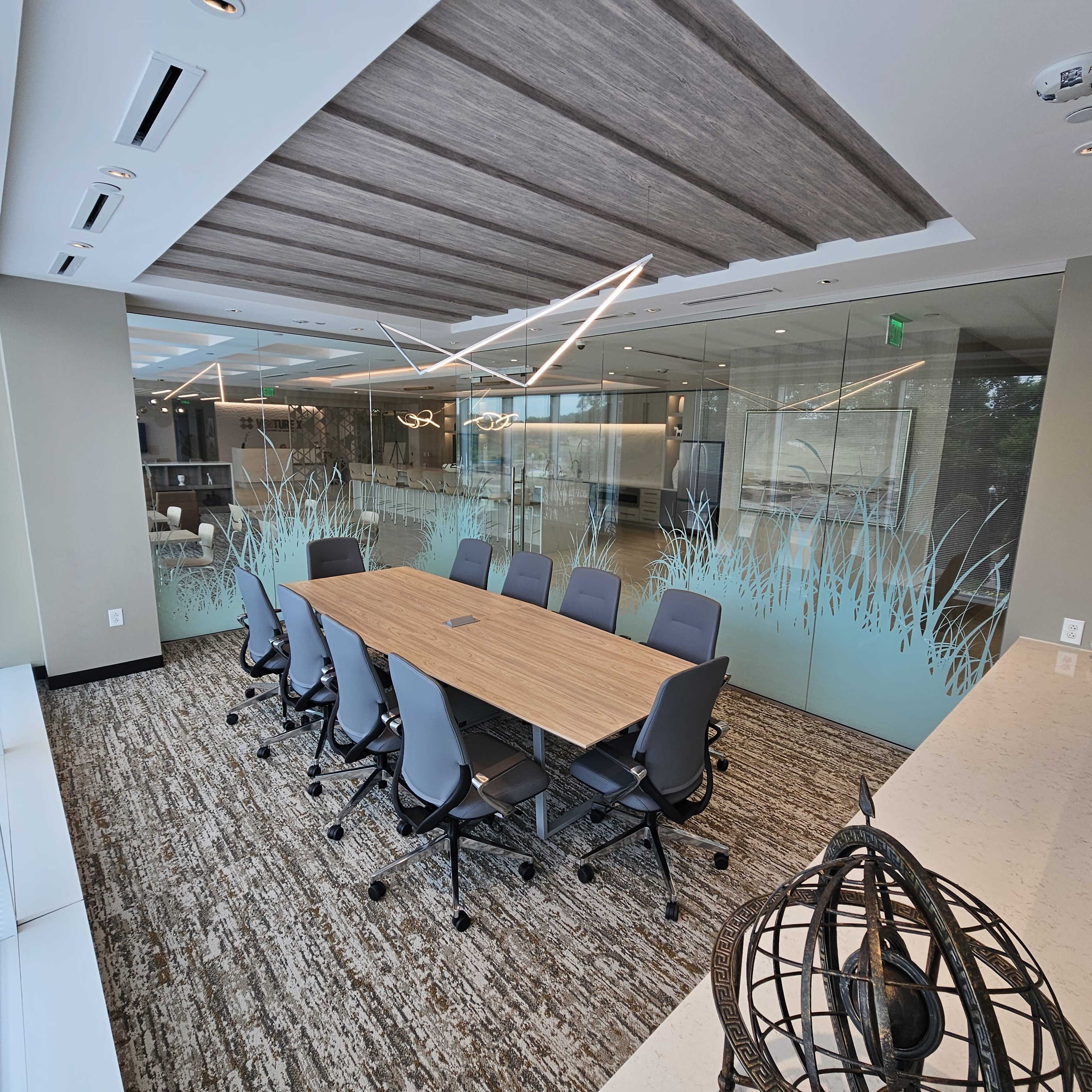 A modern conference room with a large wooden table surrounded by gray chairs, featuring glass walls and a ceiling with decorative lighting.