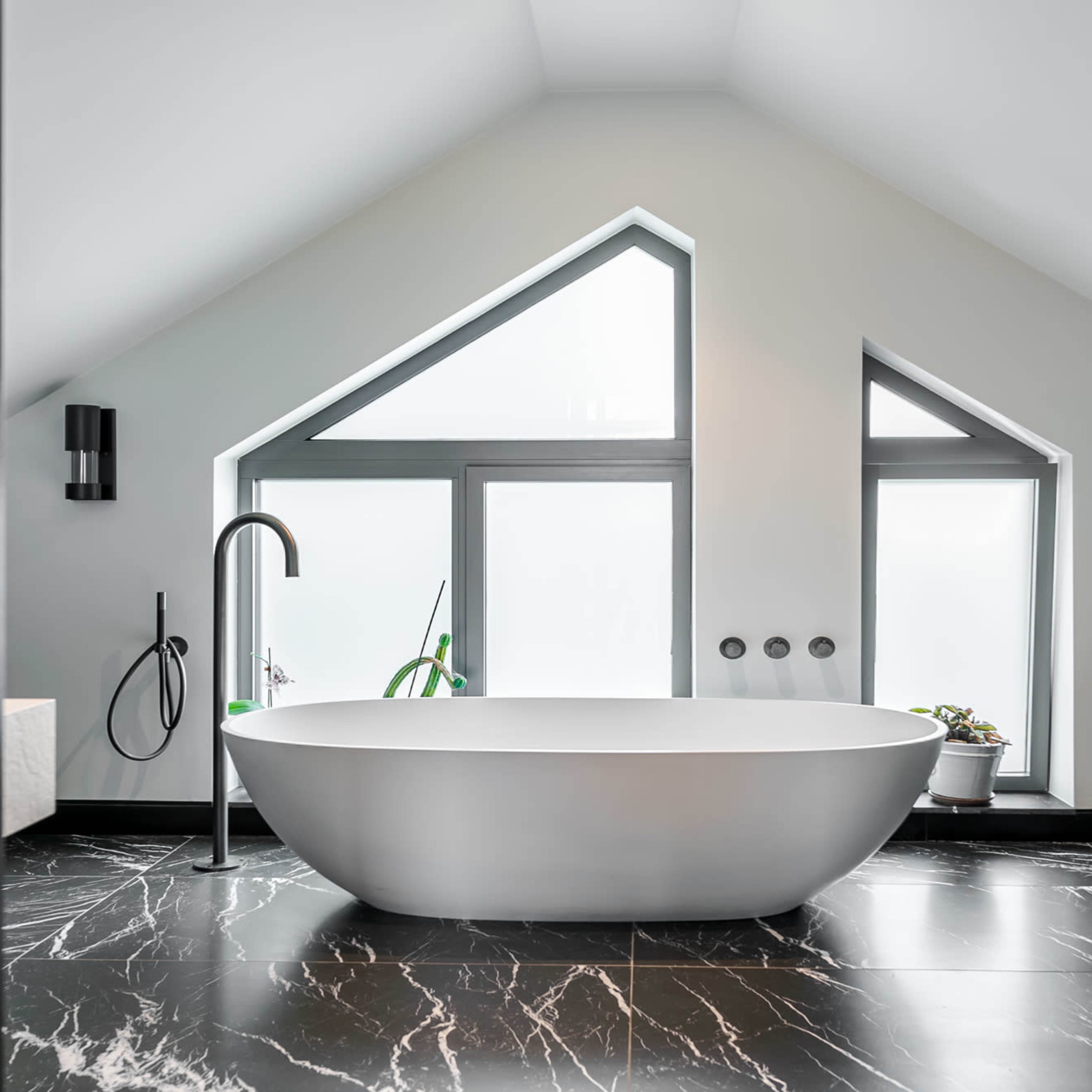 A freestanding bathtub is positioned in a modern bathroom with large triangular windows and marble flooring.