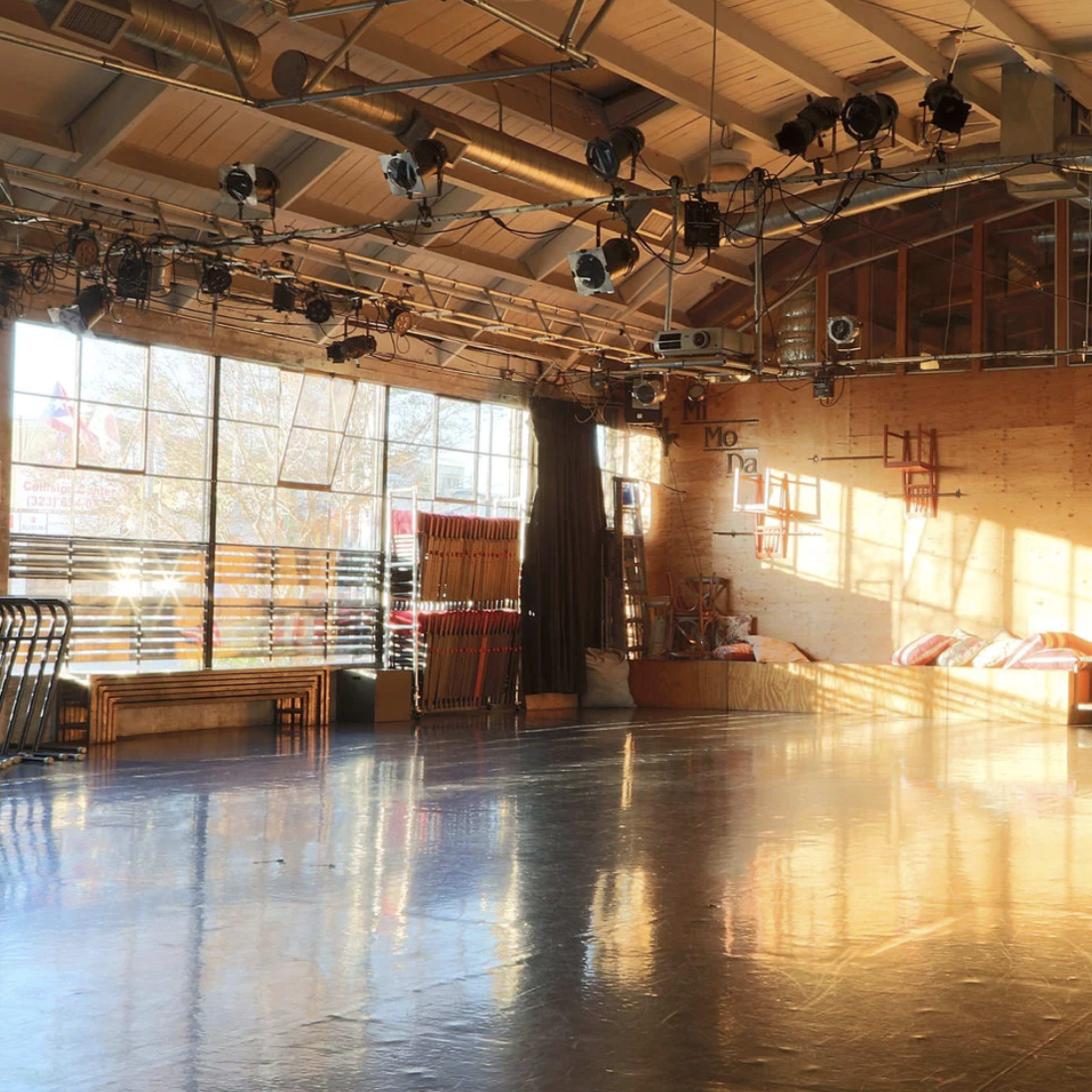 A spacious dance studio with wooden walls, large windows letting in natural light, and various lighting fixtures suspended from the ceiling.