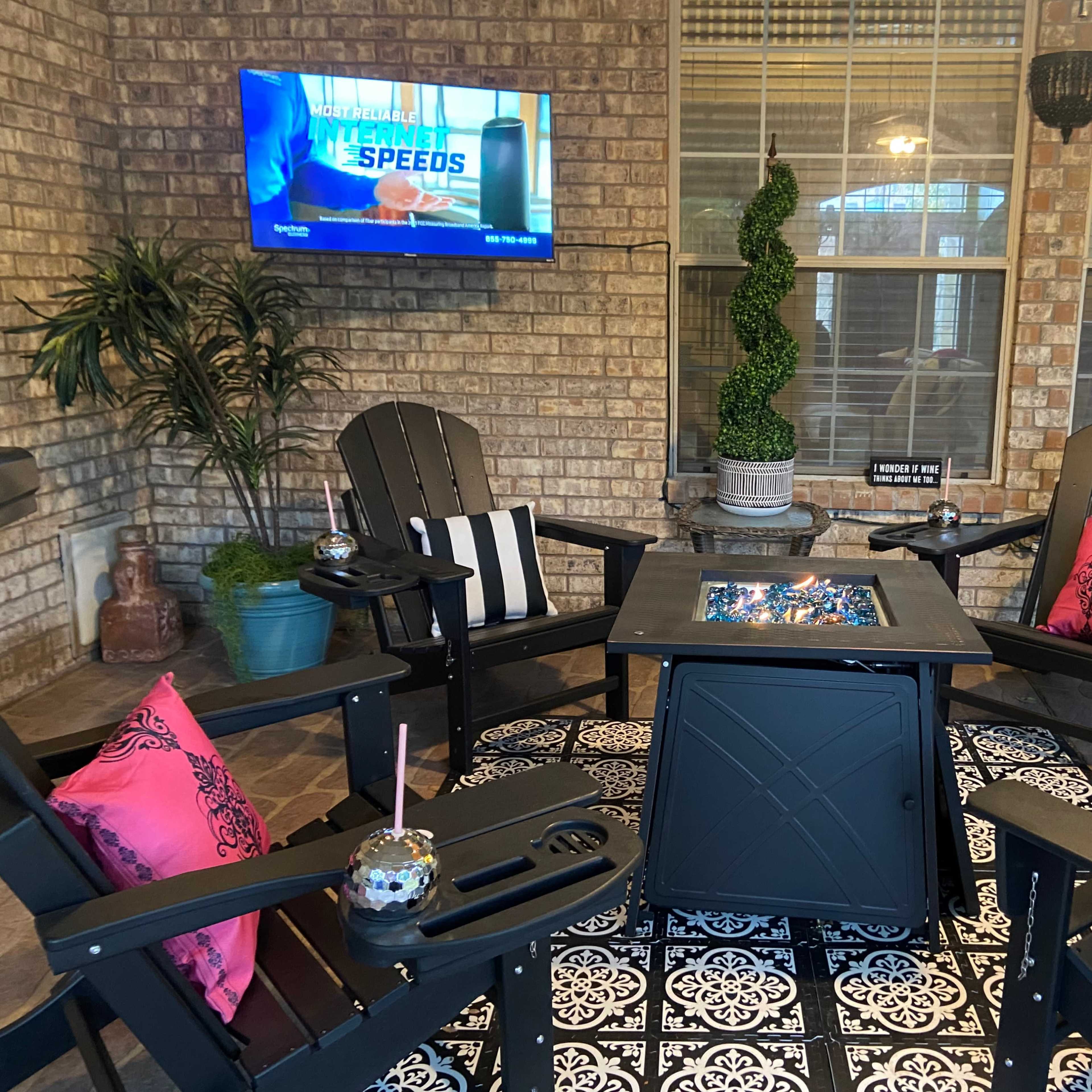A patio area features four adirondack chairs surrounding a square fire table with a television mounted on the brick wall and decorative plants in the background.