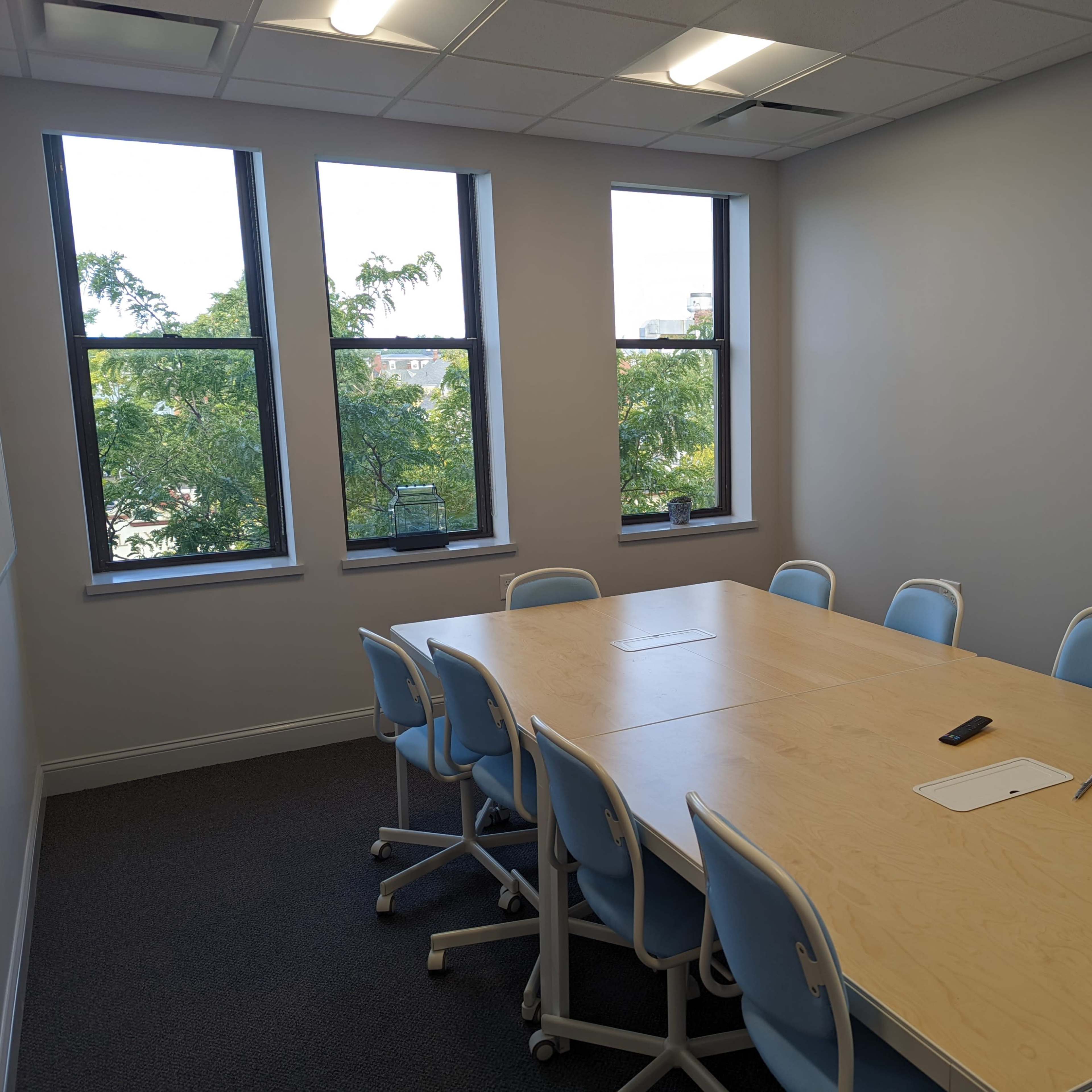 A conference room features a long wooden table surrounded by blue chairs and large windows with views of trees outside.