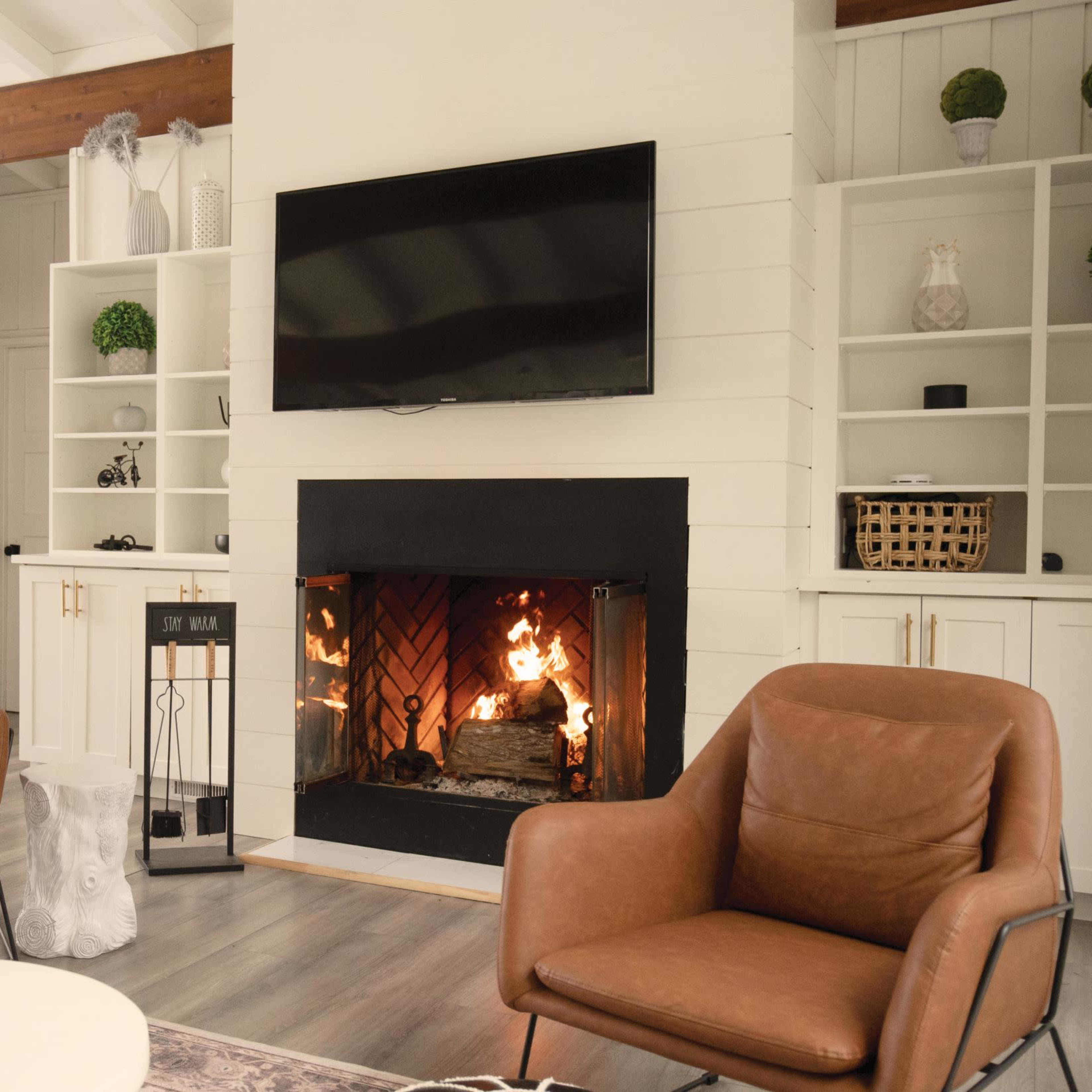 A cozy living room with a fireplace, two brown leather chairs, a round coffee table, and shelving units displaying decorative items.