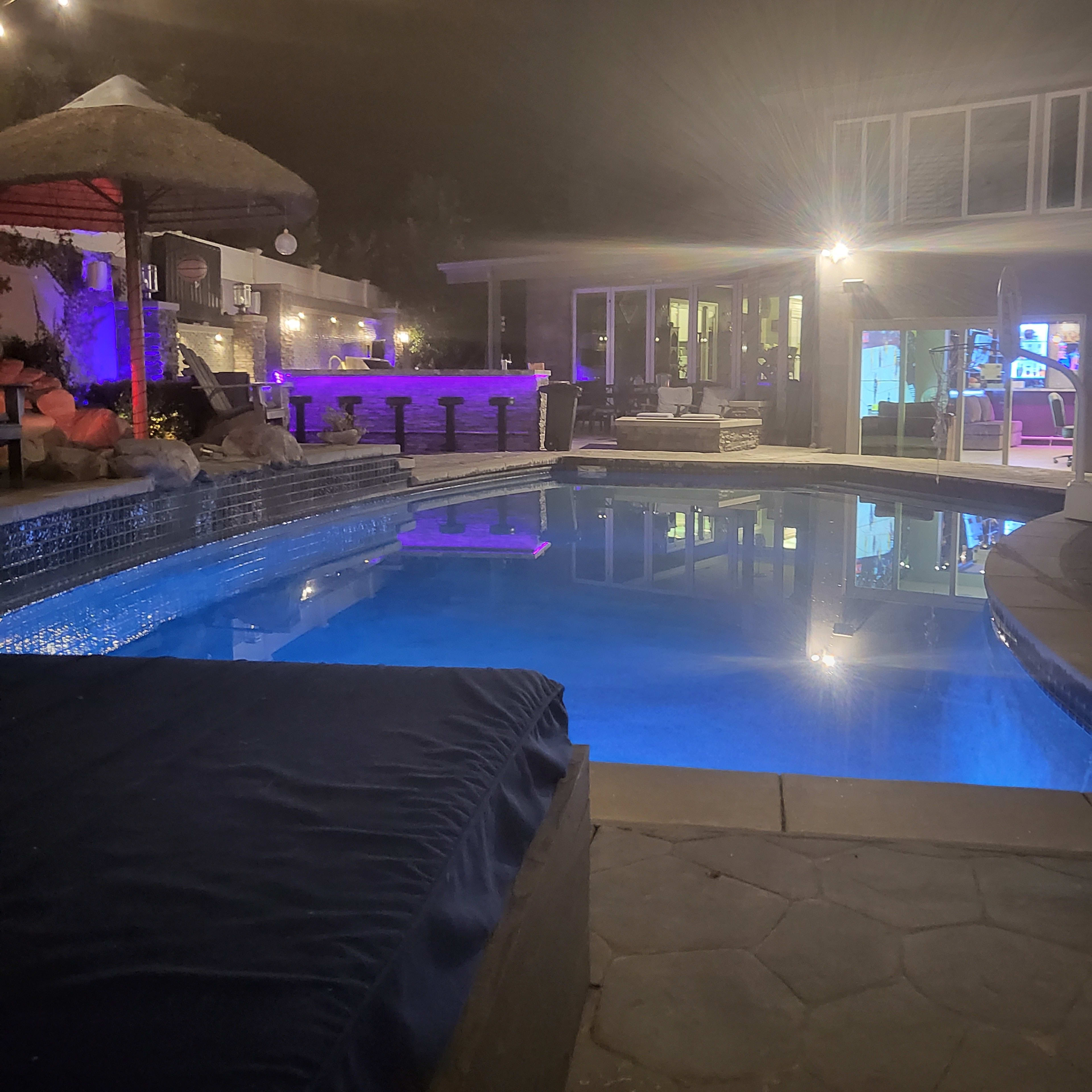 A well-lit backyard at night featuring a swimming pool with blue water, a thatched umbrella, and an outdoor seating area.