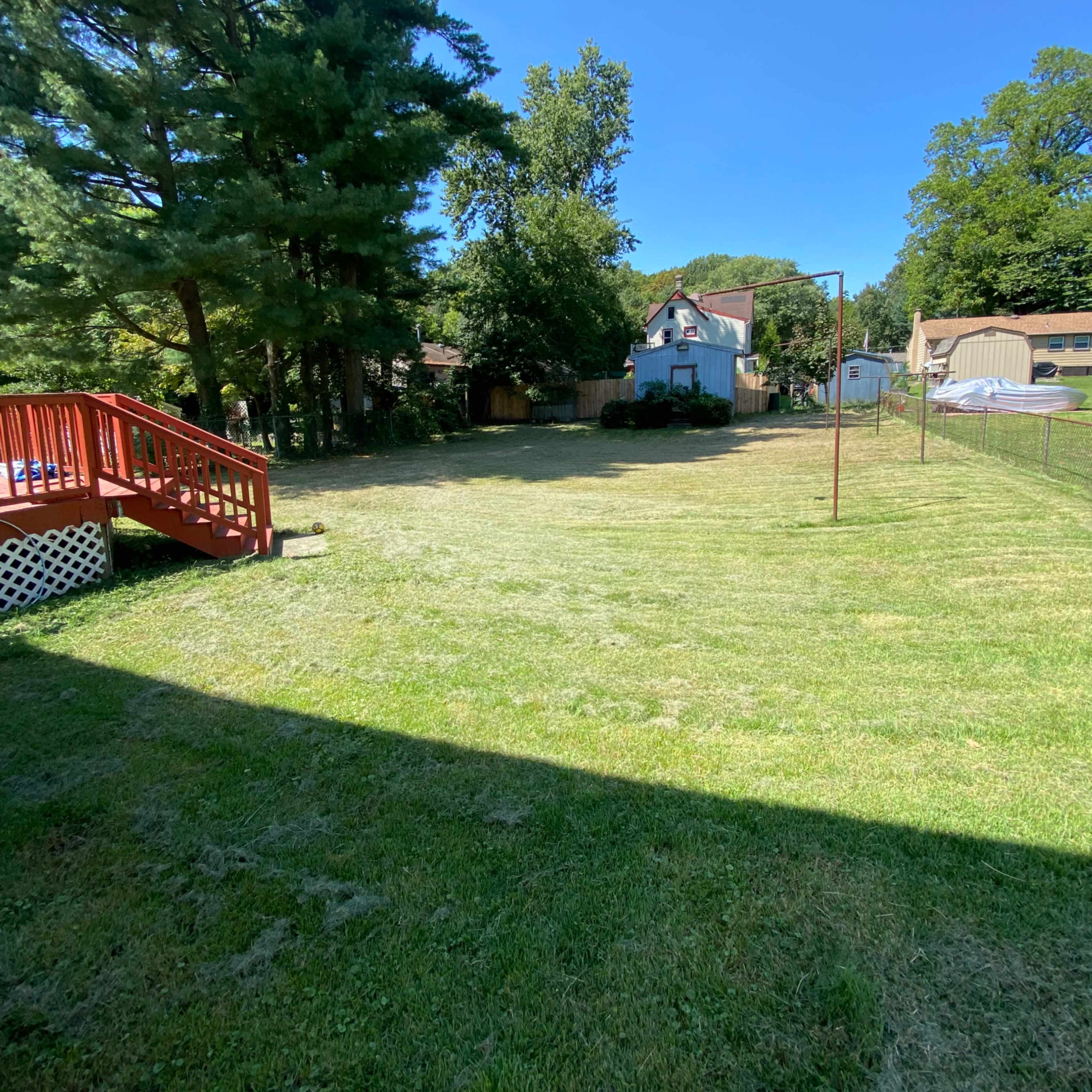 A grassy backyard with a wooden deck, several trees, and a house in the background.