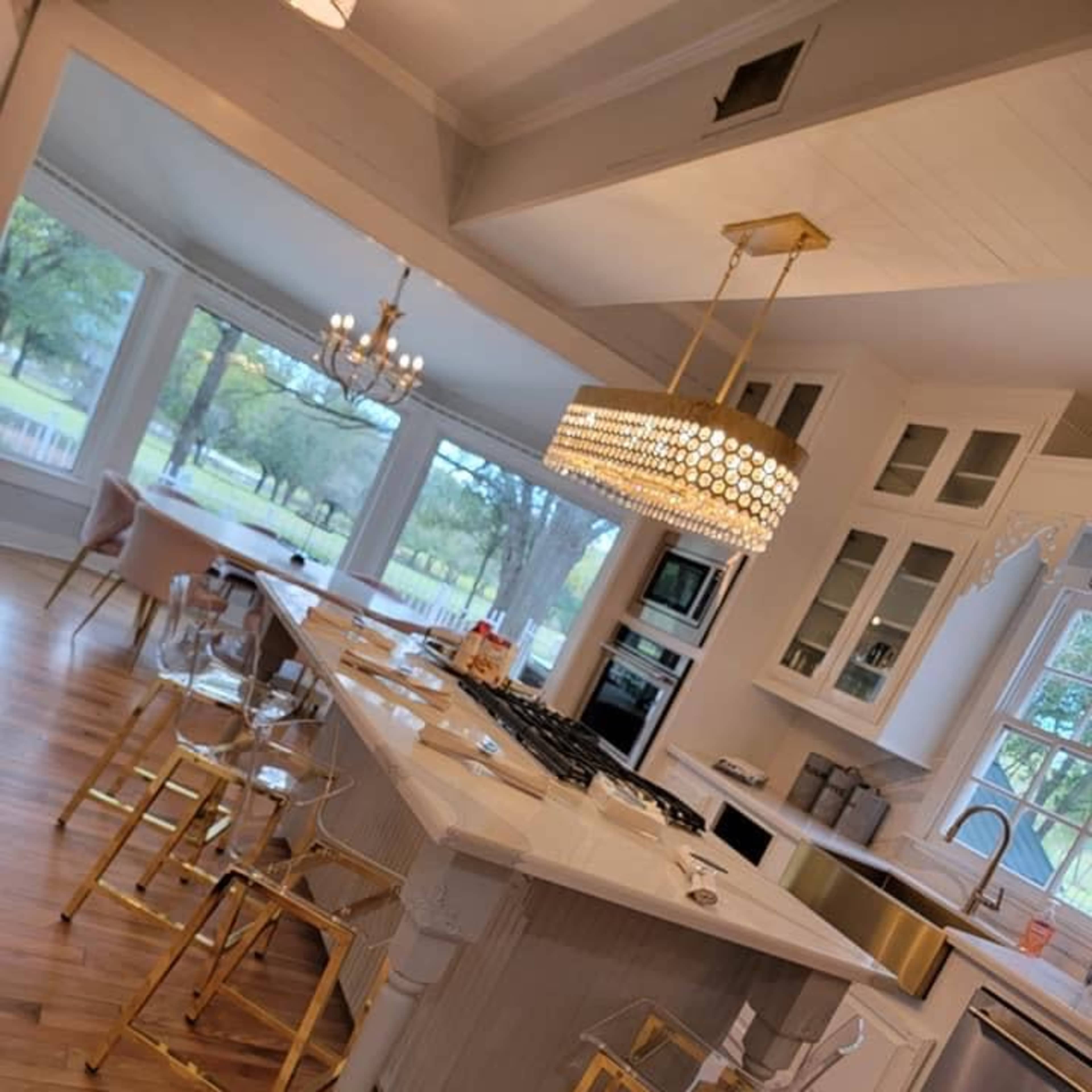 A modern kitchen features a large island with gold-accented bar stools, pendant lighting, and a view of the outdoors through large windows.