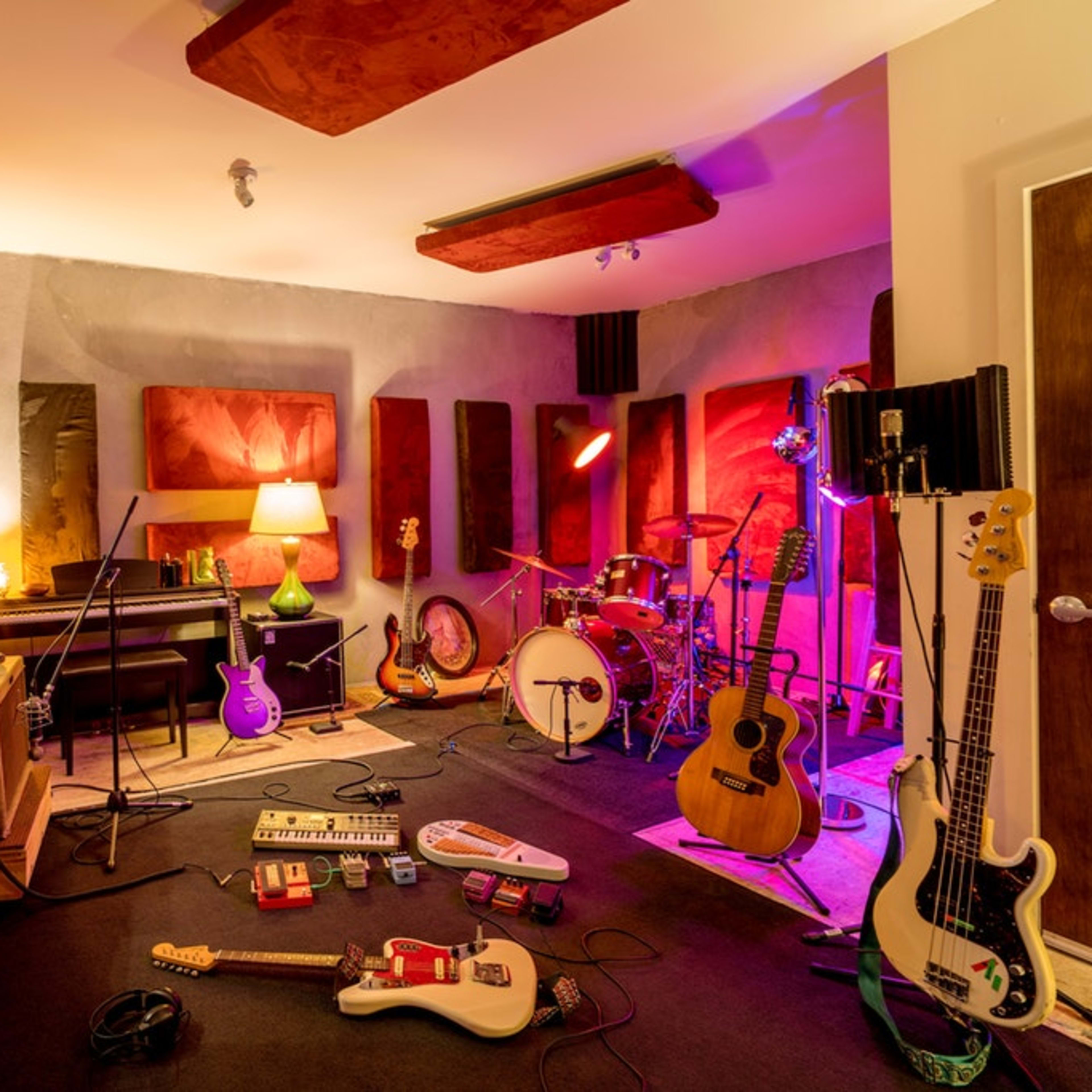 A music recording studio features various instruments scattered on the floor, soundproofing panels on the walls, and a drum set set up in the corner.