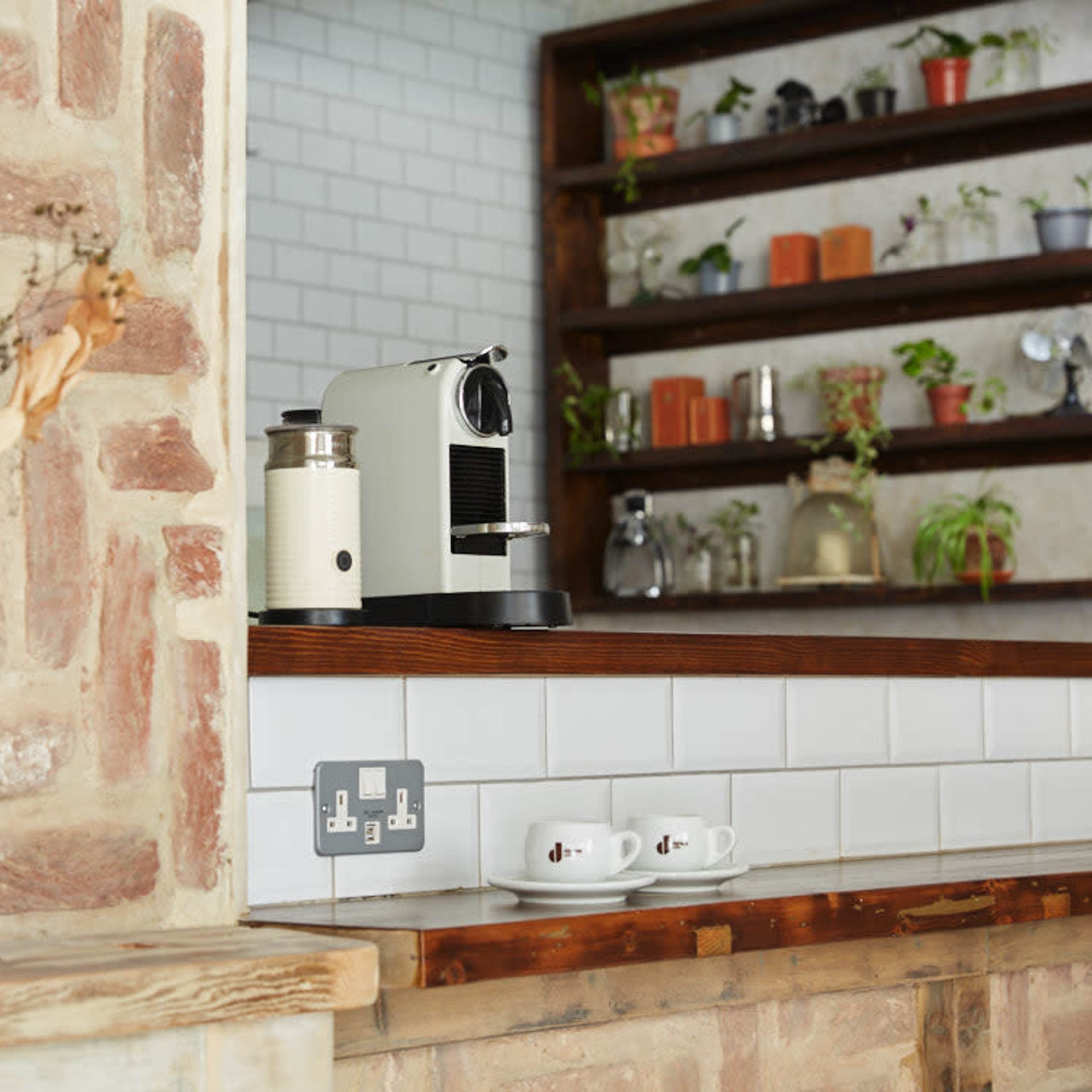 A kitchen with a coffee machine on a wooden countertop, a vase of dried flowers, and shelves containing plants and decorative items.