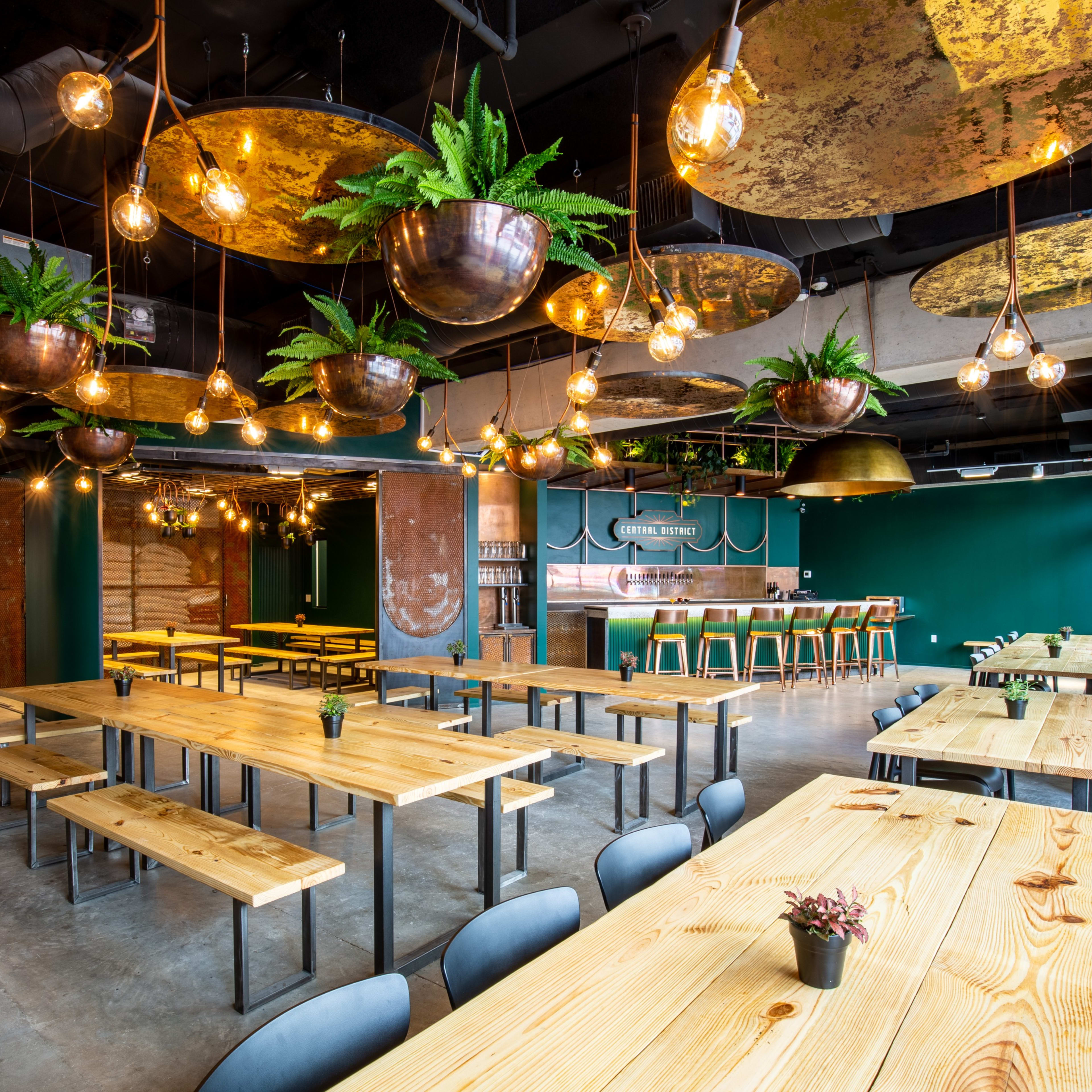 A modern restaurant interior features long wooden tables, black chairs, and hanging light fixtures with greenery against a backdrop of teal and metallic decor.