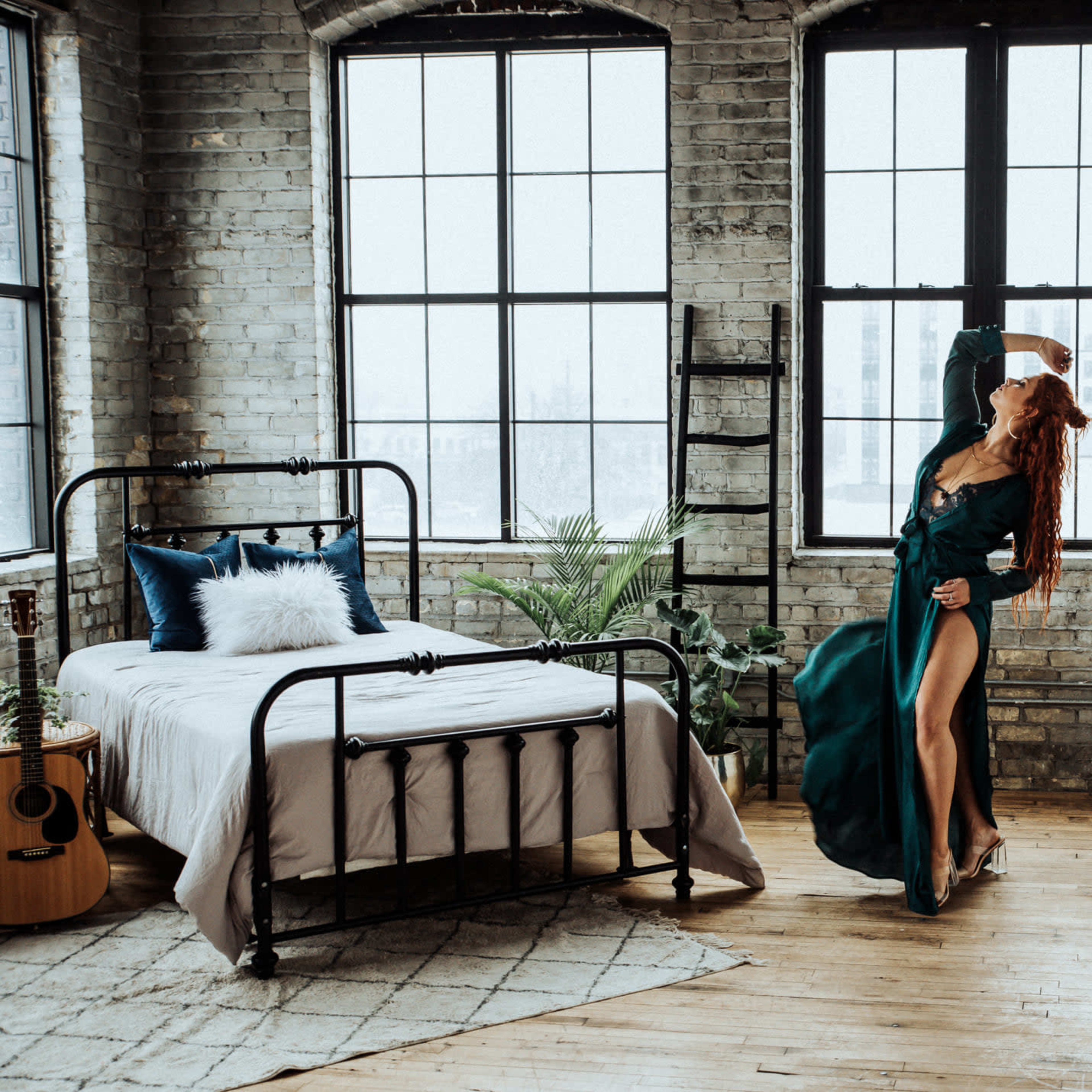 A person in a green dress poses near a large window in an industrial-style bedroom with a bed, guitar, and plants.