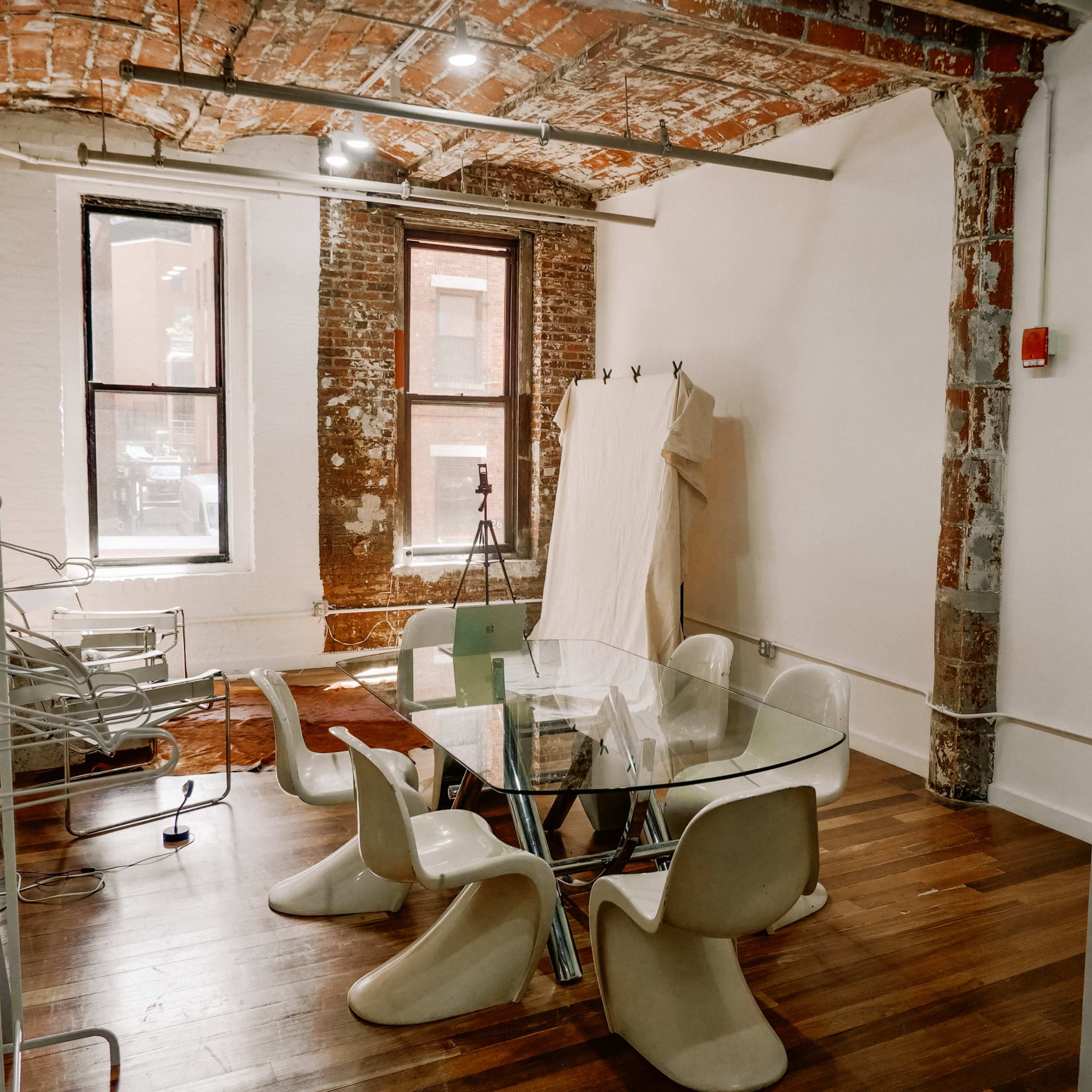 A modern meeting room featuring a glass table surrounded by sculptural chairs, with exposed brick walls and large windows.