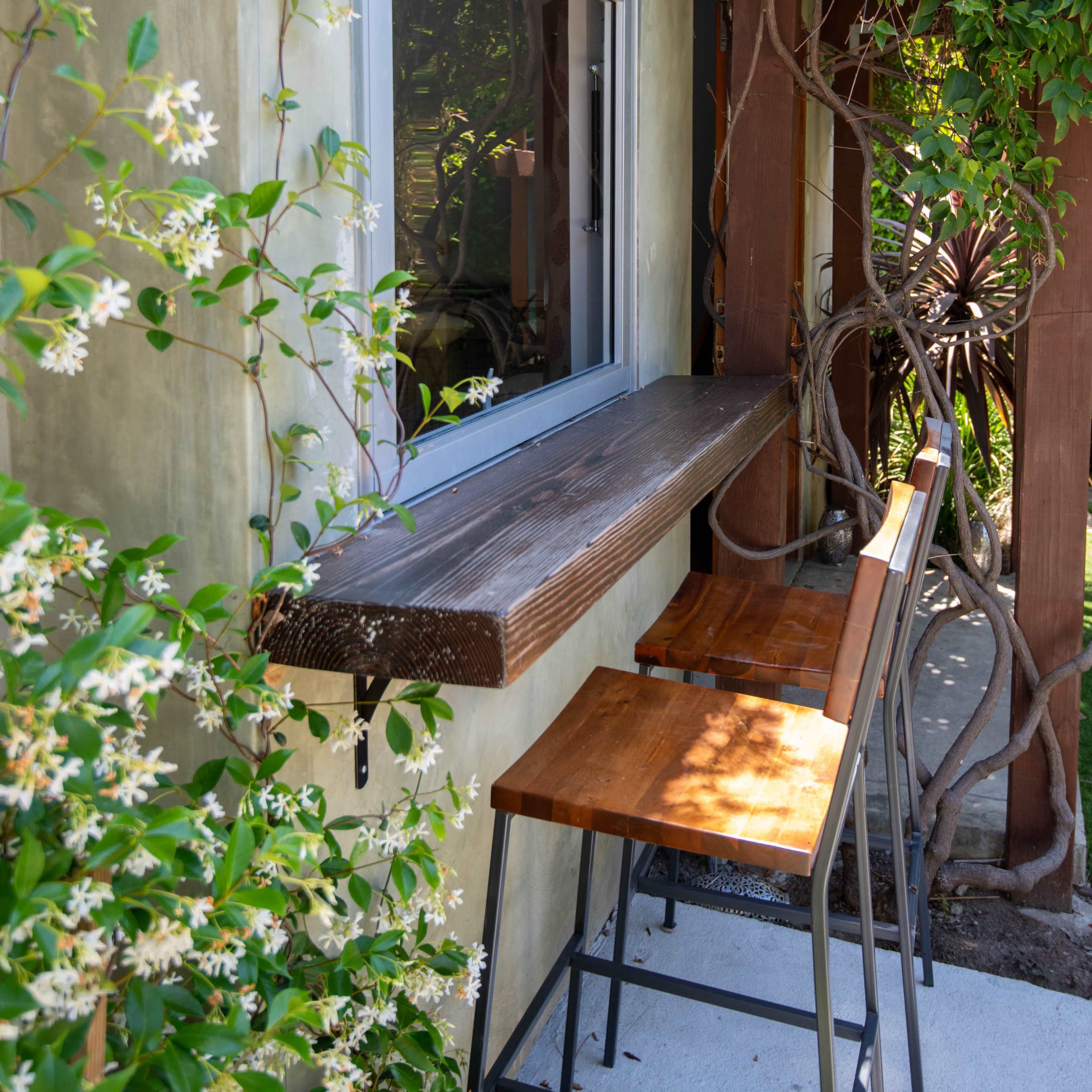 A wooden countertop with two metal-framed stools is positioned beside a window surrounded by green vines and flowers.