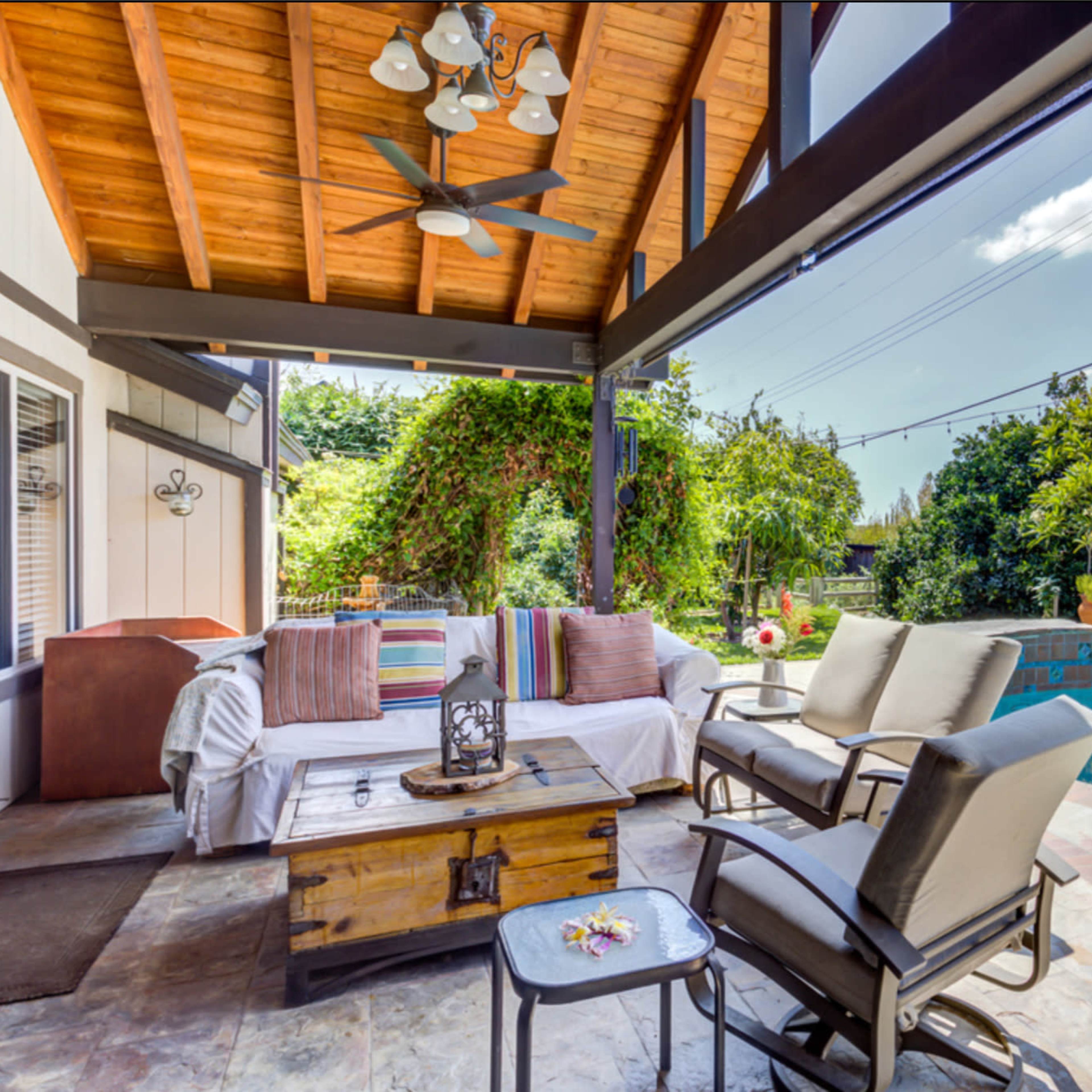 A covered patio features a seating area with a coffee table and a view of a swimming pool surrounded by greenery.