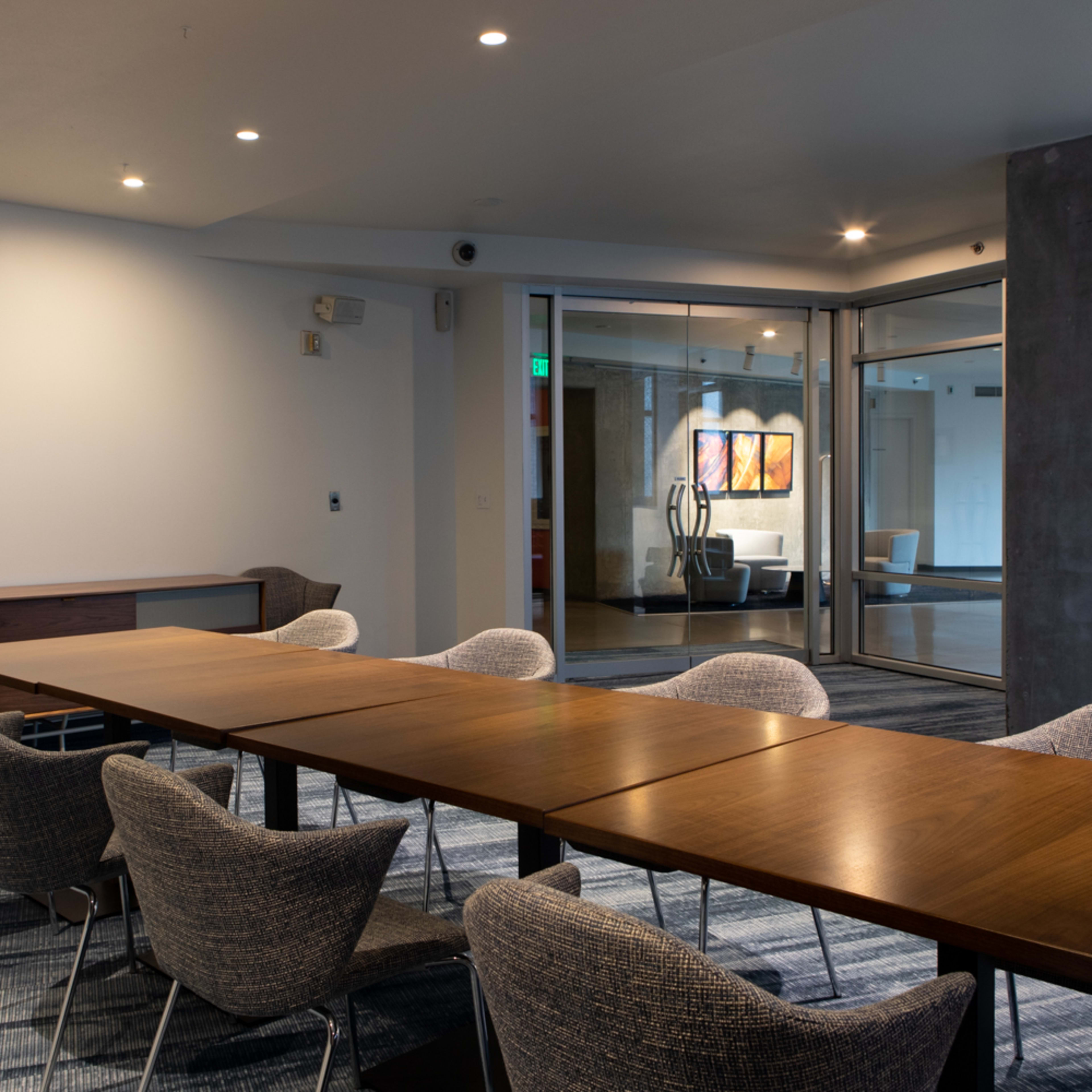 A long wooden table surrounded by eight chairs is set in a well-lit conference room, with a glass door leading to an adjoining area.