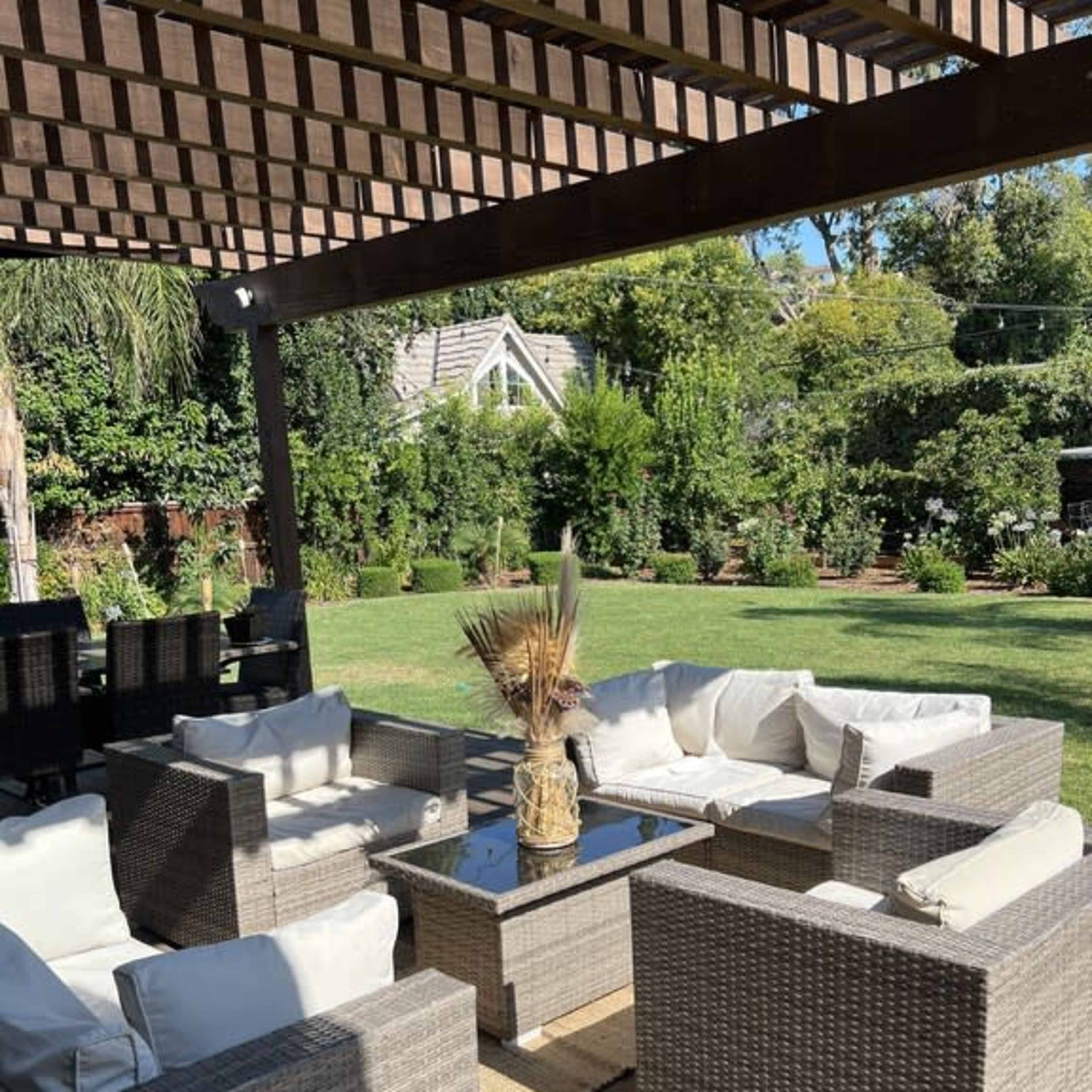A patio seating area features wicker furniture with white cushions arranged around a glass-topped table, set beneath a wooden pergola in a landscaped yard.