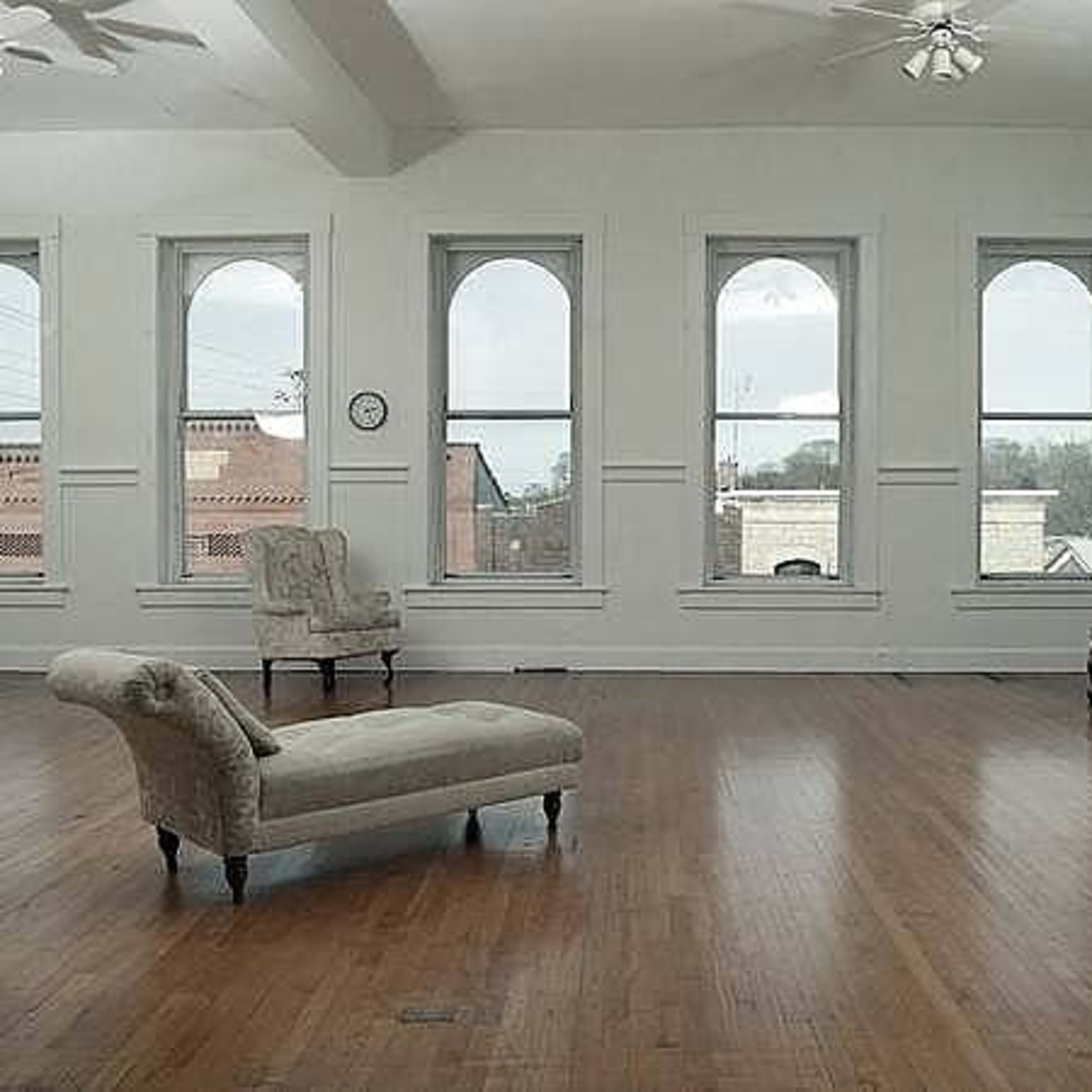 A spacious, light-filled room with large arched windows, hardwood flooring, and two upholstered chairs placed against the walls.