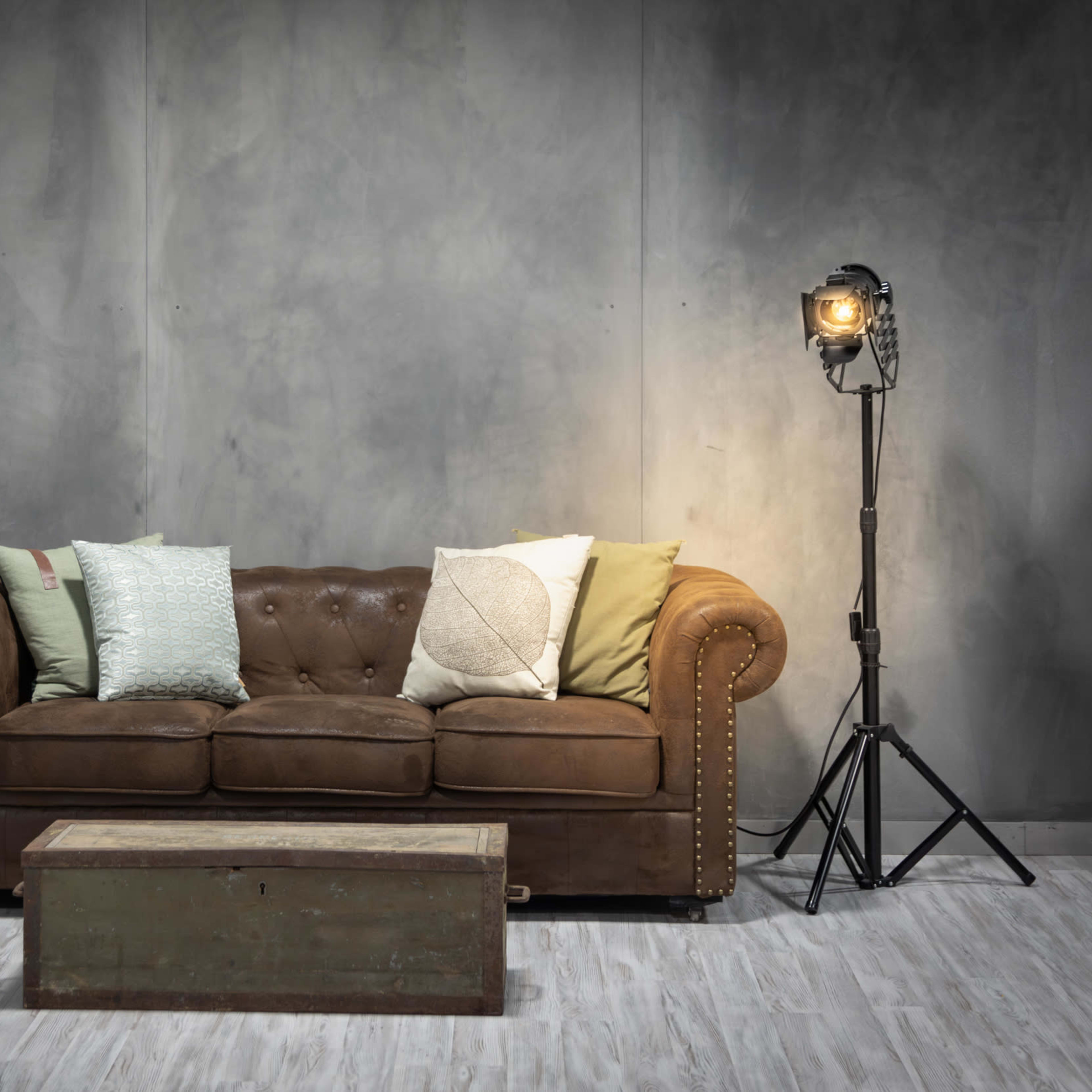 A brown leather sofa with colorful pillows, a wooden coffee table, a green locker, and a standing light on a gray backdrop.