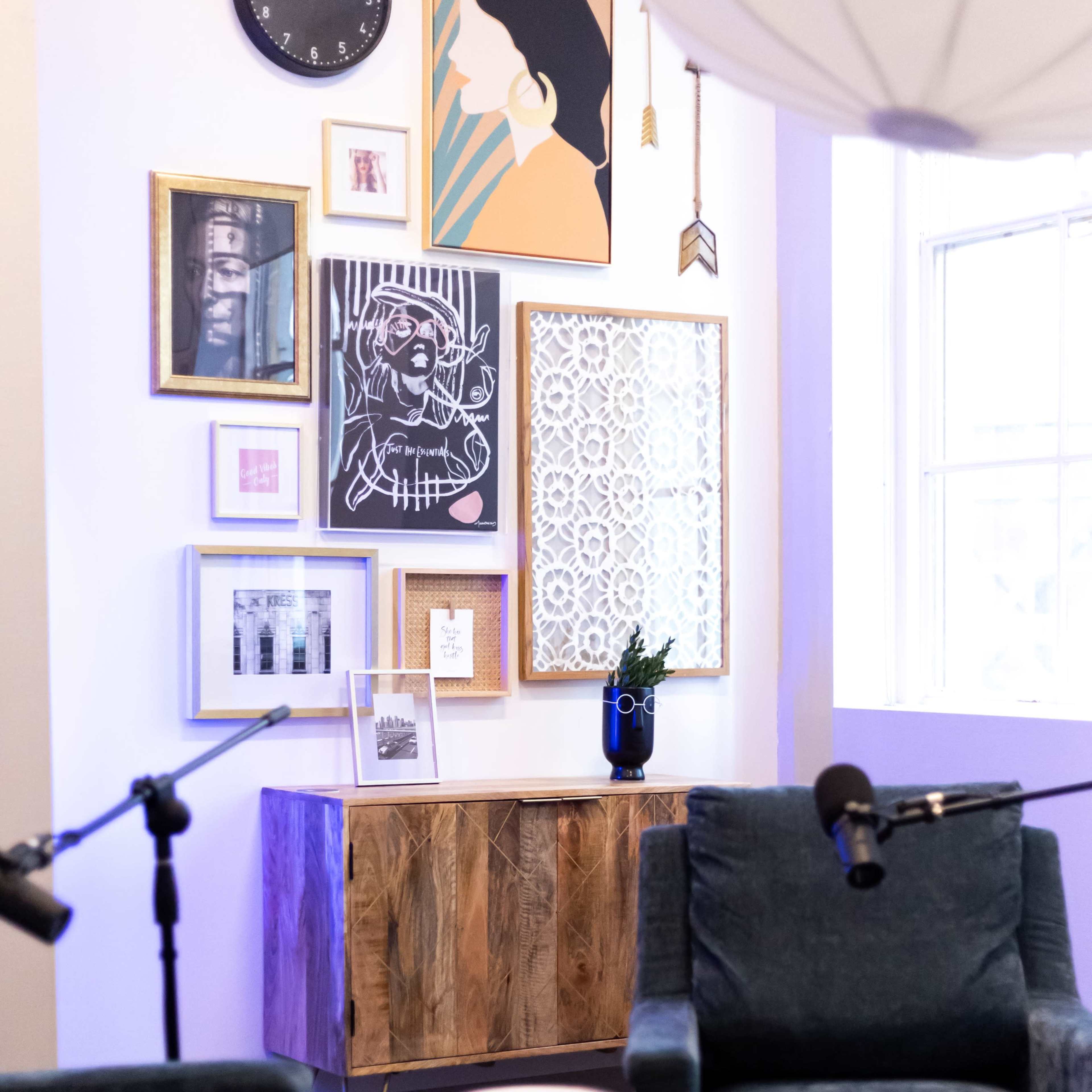 A corner of a room featuring a wooden cabinet, a variety of framed artworks on the wall, a clock, a plant, and two upholstered chairs with a small table in between.
