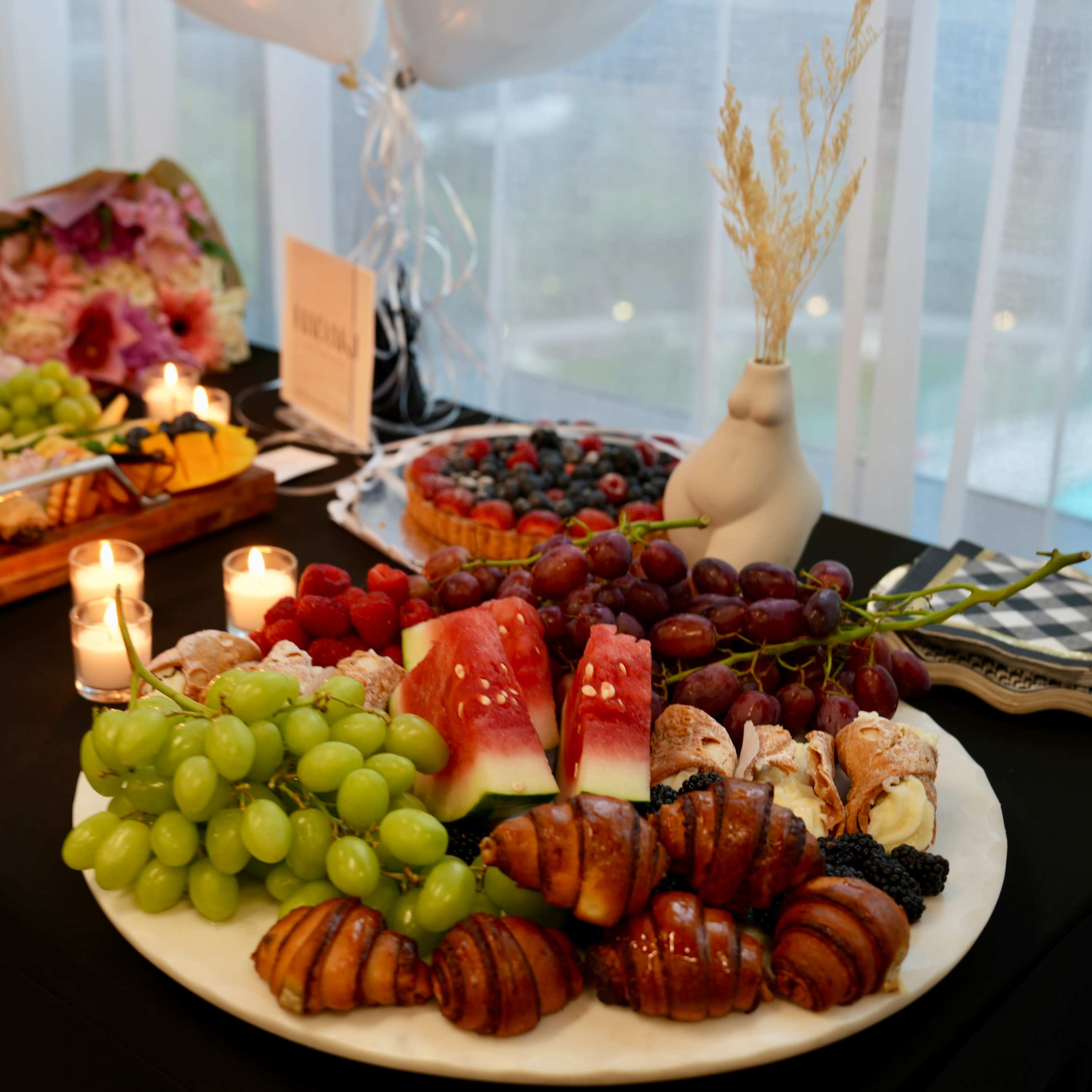 A variety of fruits, pastries, and a charcuterie board are displayed on a table adorned with candles and decorative elements.