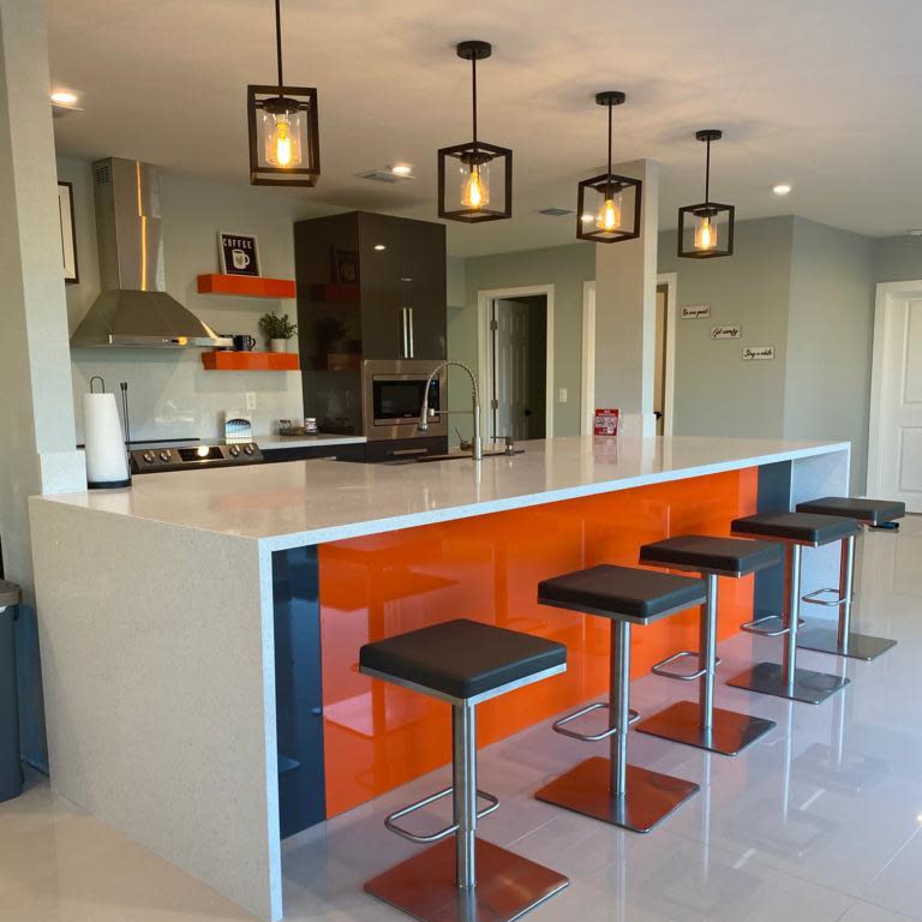 A modern kitchen features a central island with an orange accent and five black bar stools arranged around it.