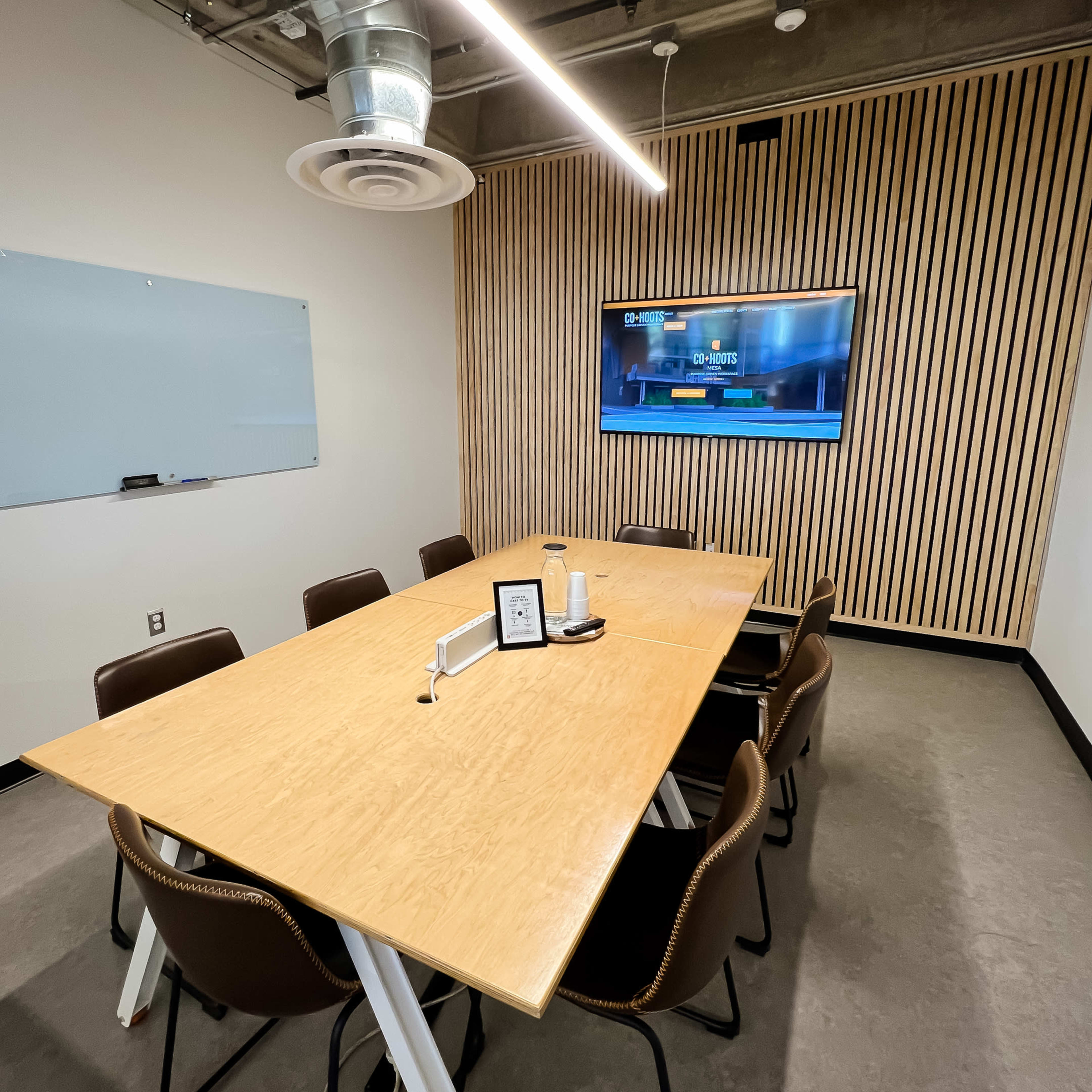 A modern conference room features a long wooden table with chairs, a wall-mounted TV, a whiteboard, and wooden paneling.