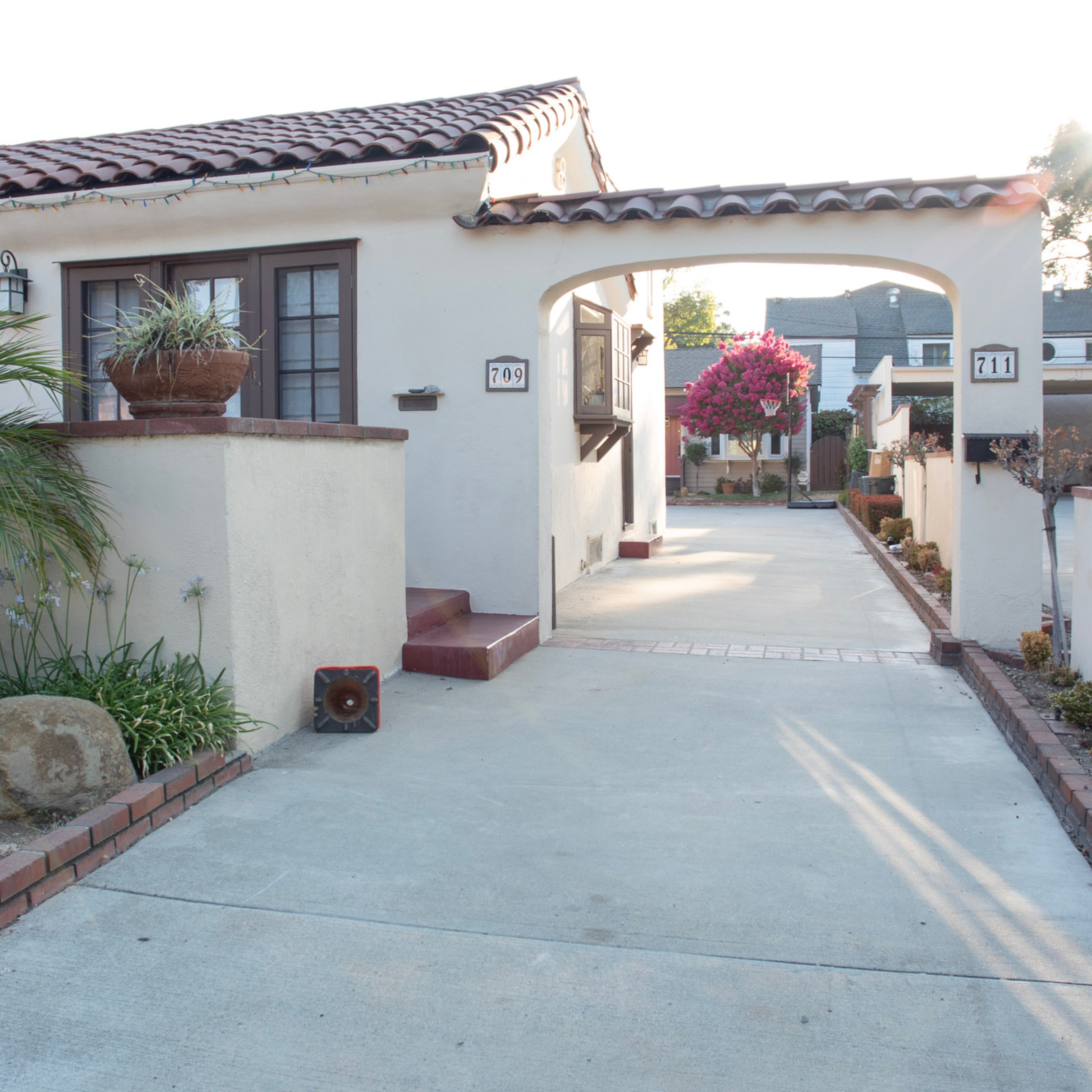 A quiet residential street with houses on either side, lined by a paved driveway leading to two mailbox posts.