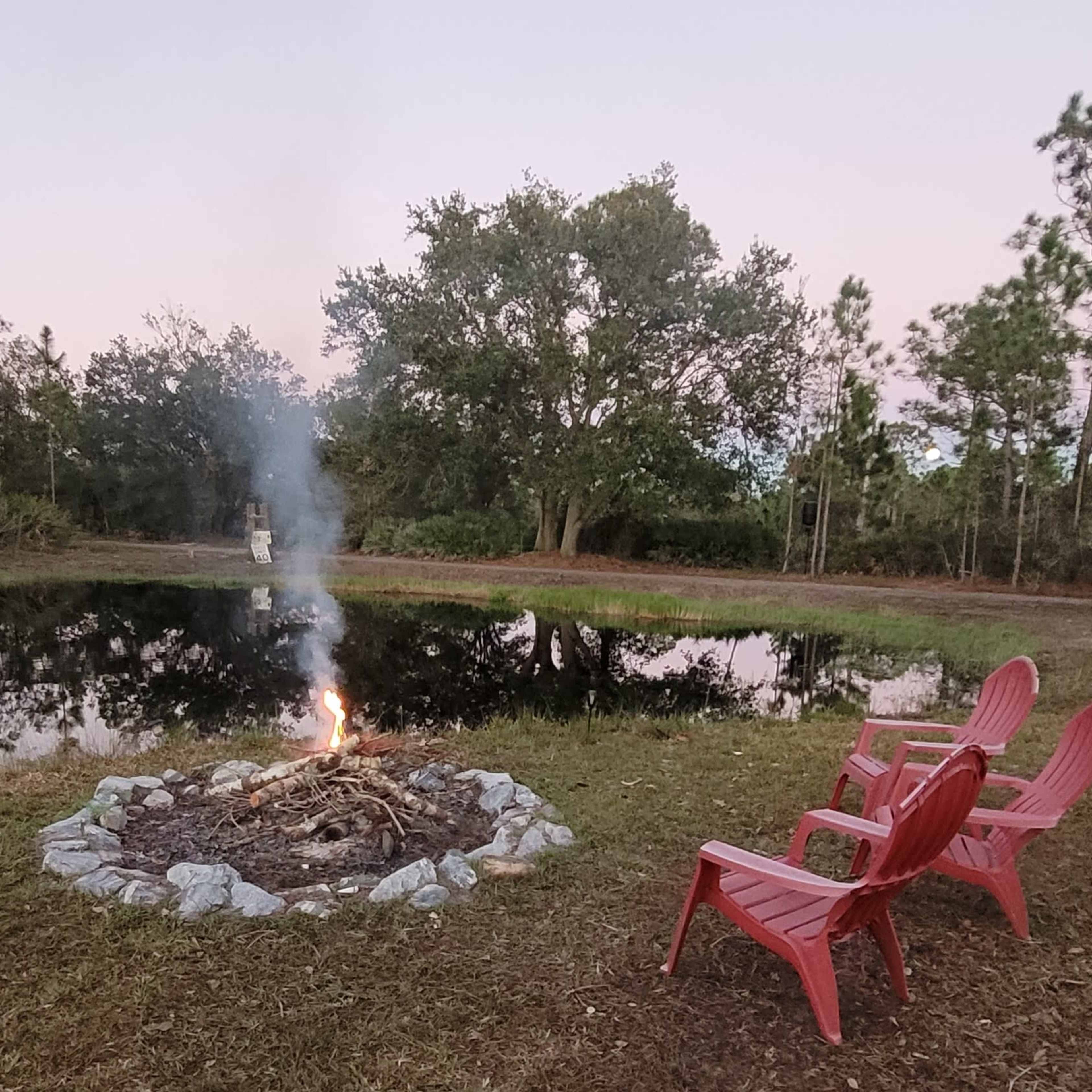 A small campfire burns in a stone circle by a pond, with two red chairs positioned nearby.