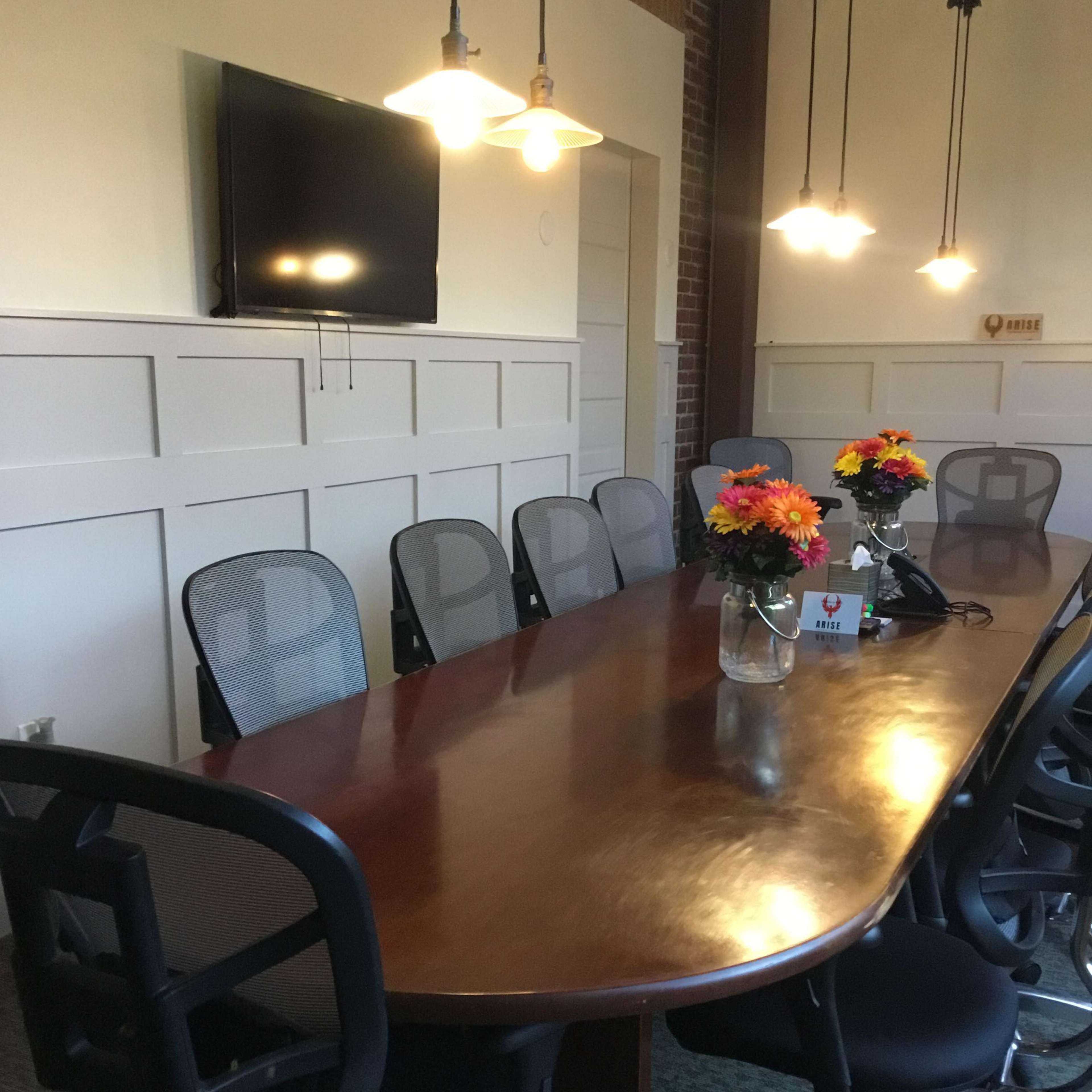 A large, oval conference table with eight ergonomic chairs is set up in a meeting room and is adorned with vases of flowers, while a television is mounted on the wall.