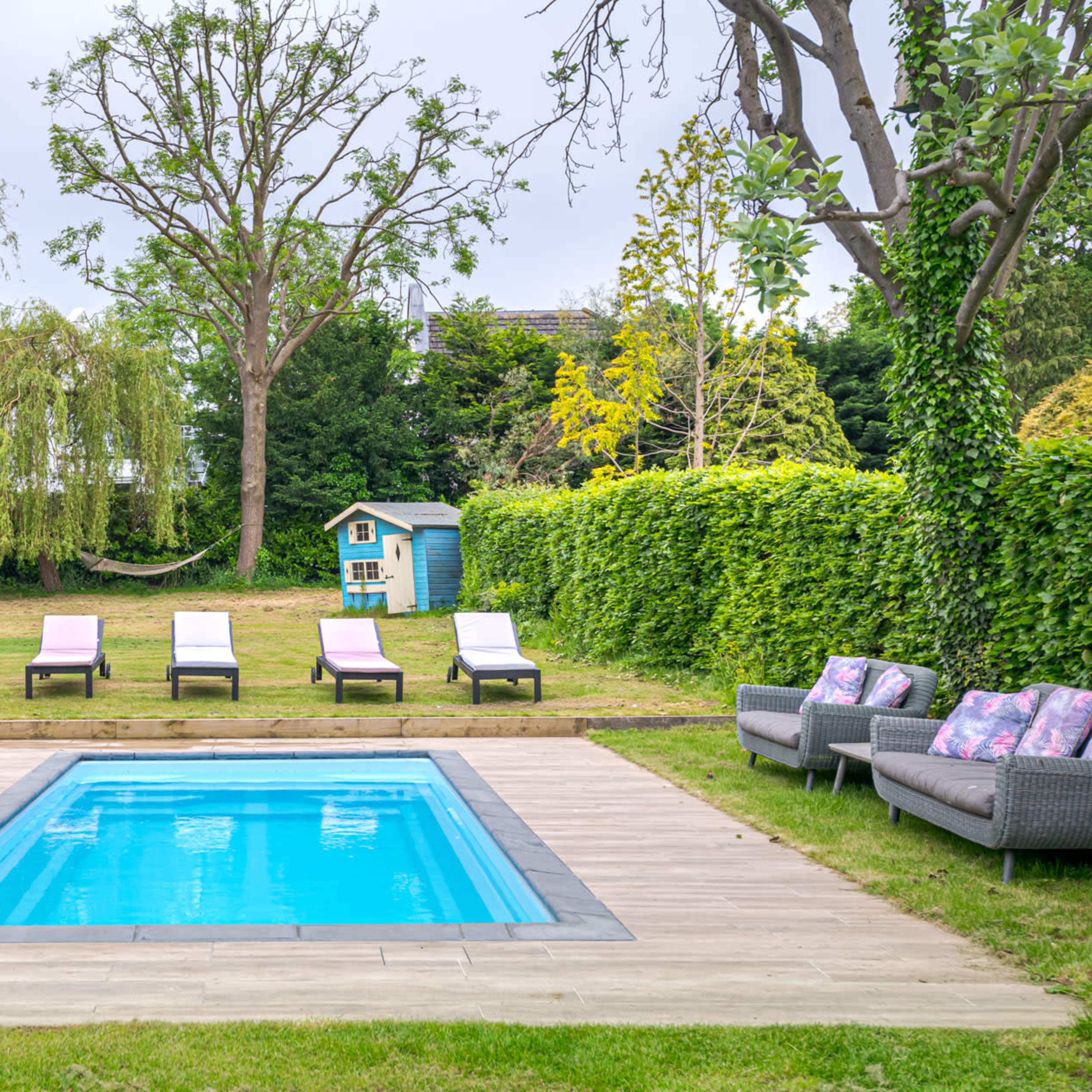 A backyard featuring a rectangular swimming pool surrounded by wooden decking, with loungers arranged nearby and a play set in the background.