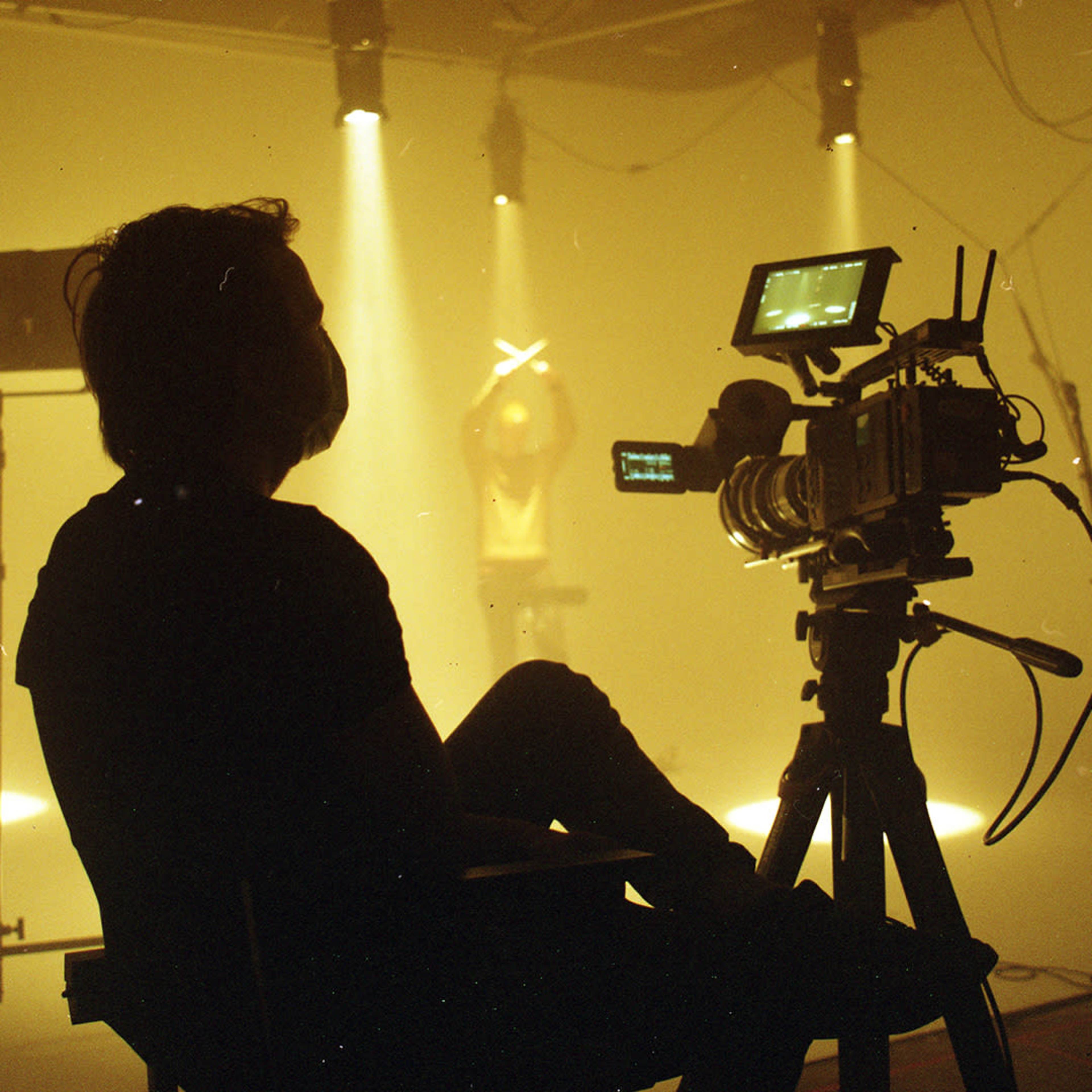 A silhouette of a person sits on a chair in front of a camera while another figure is illuminated in the background under spotlights.