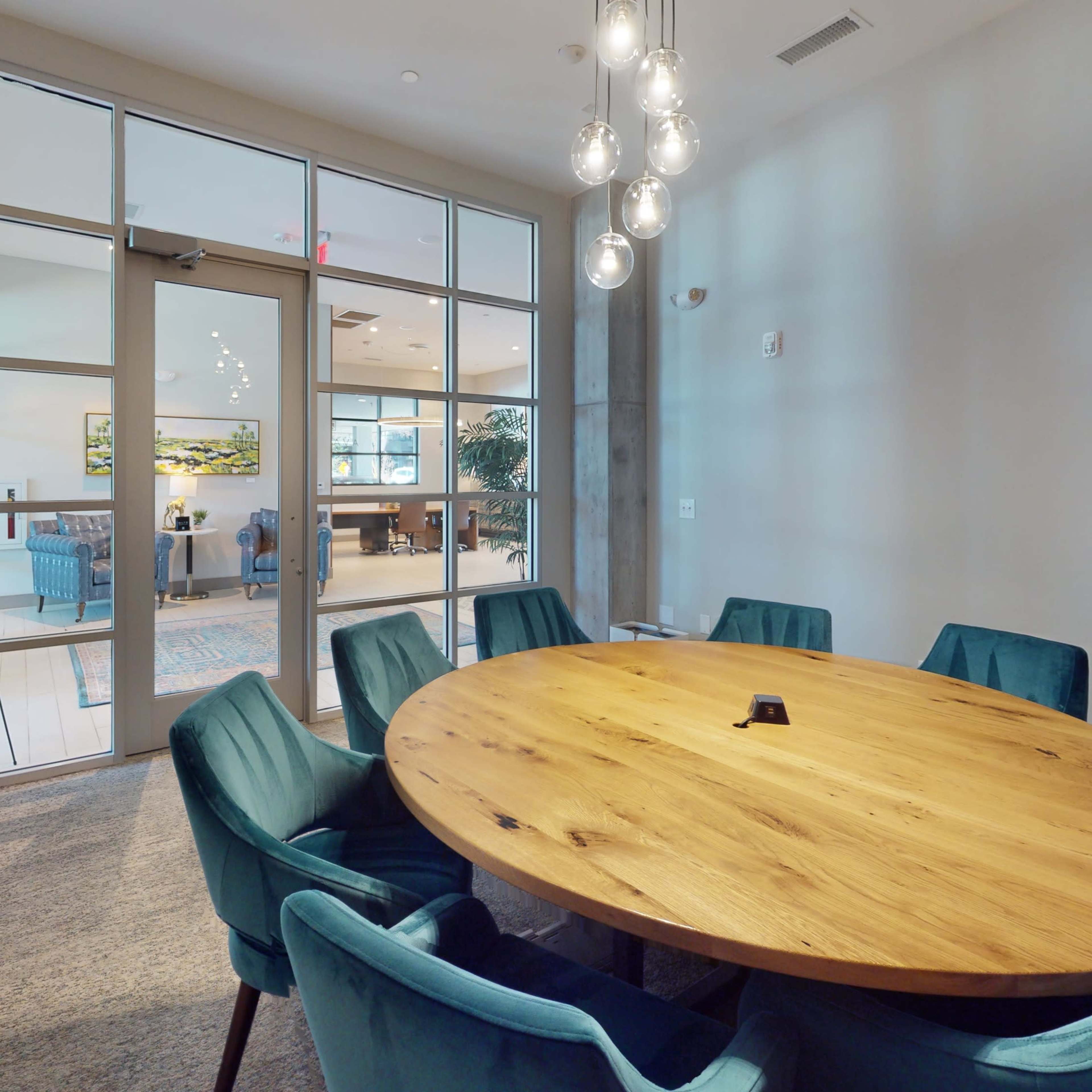 A round wooden table surrounded by teal chairs is positioned in a well-lit conference room with large windows and contemporary decor.