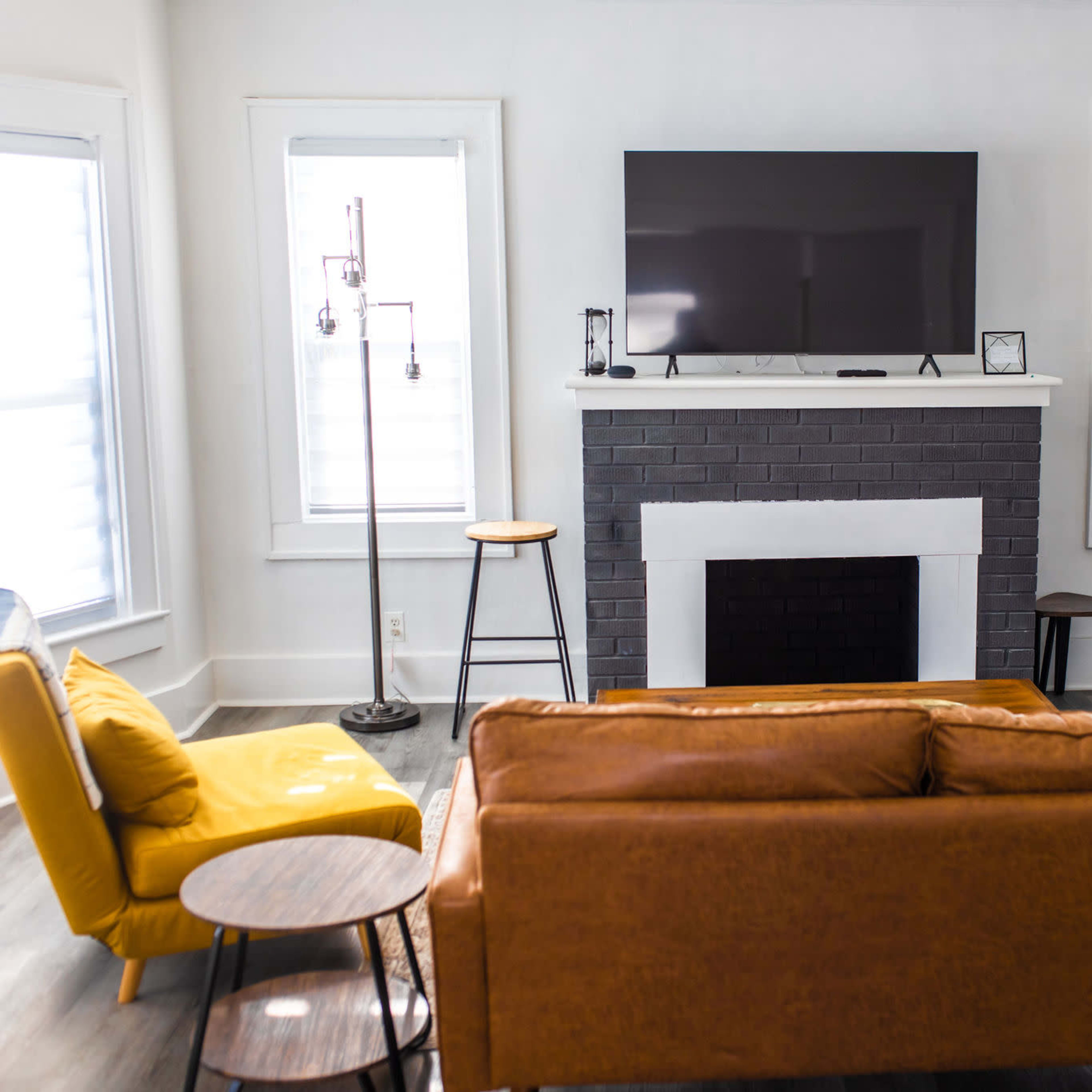 The living room features a brown leather sofa, a yellow armchair, a flat-screen TV mounted above a black brick fireplace, and several wooden stools arranged around the space.