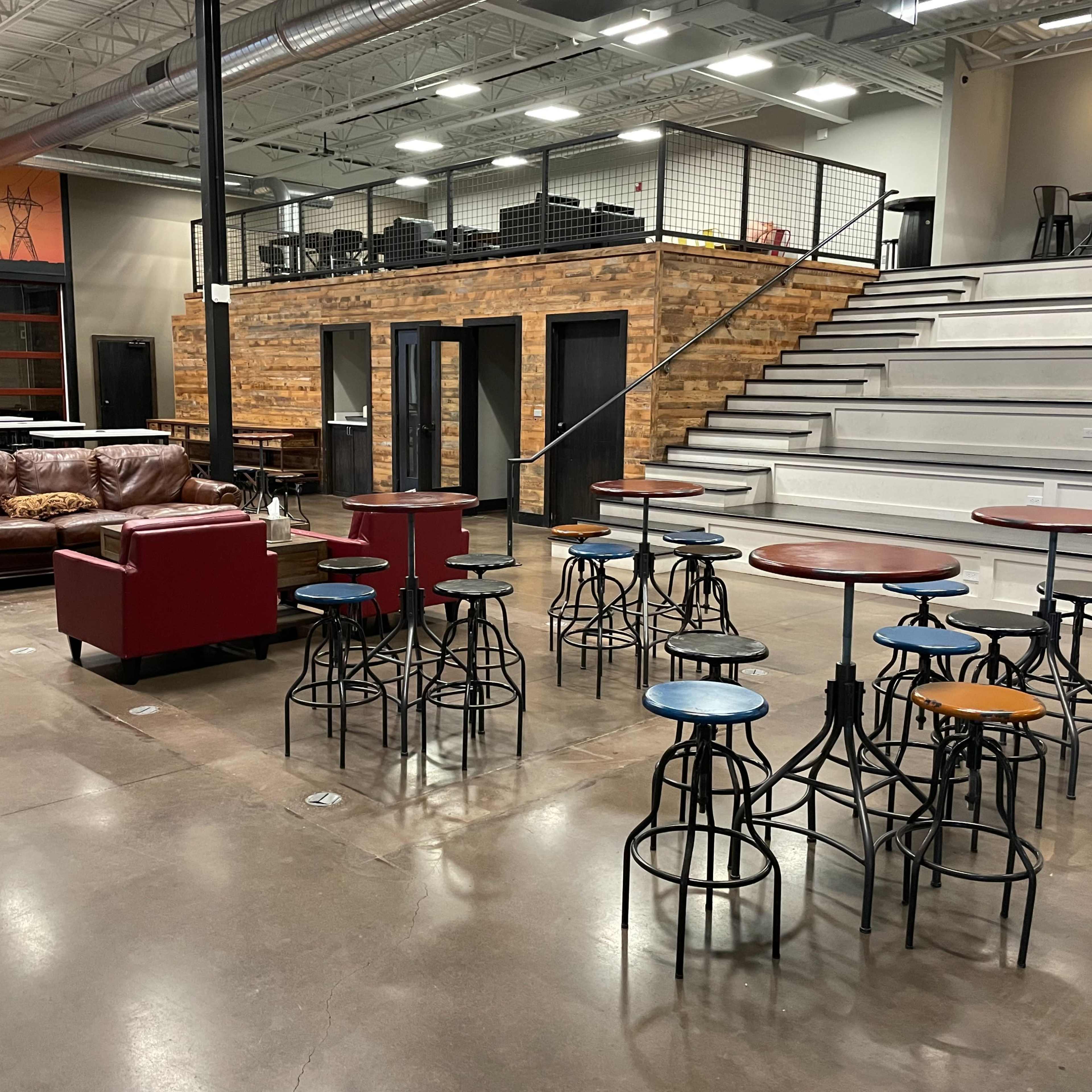 A spacious indoor area featuring a mix of seating options, including a brown leather sofa and several high tables surrounded by stools, with a multi-level seating arrangement in the background.