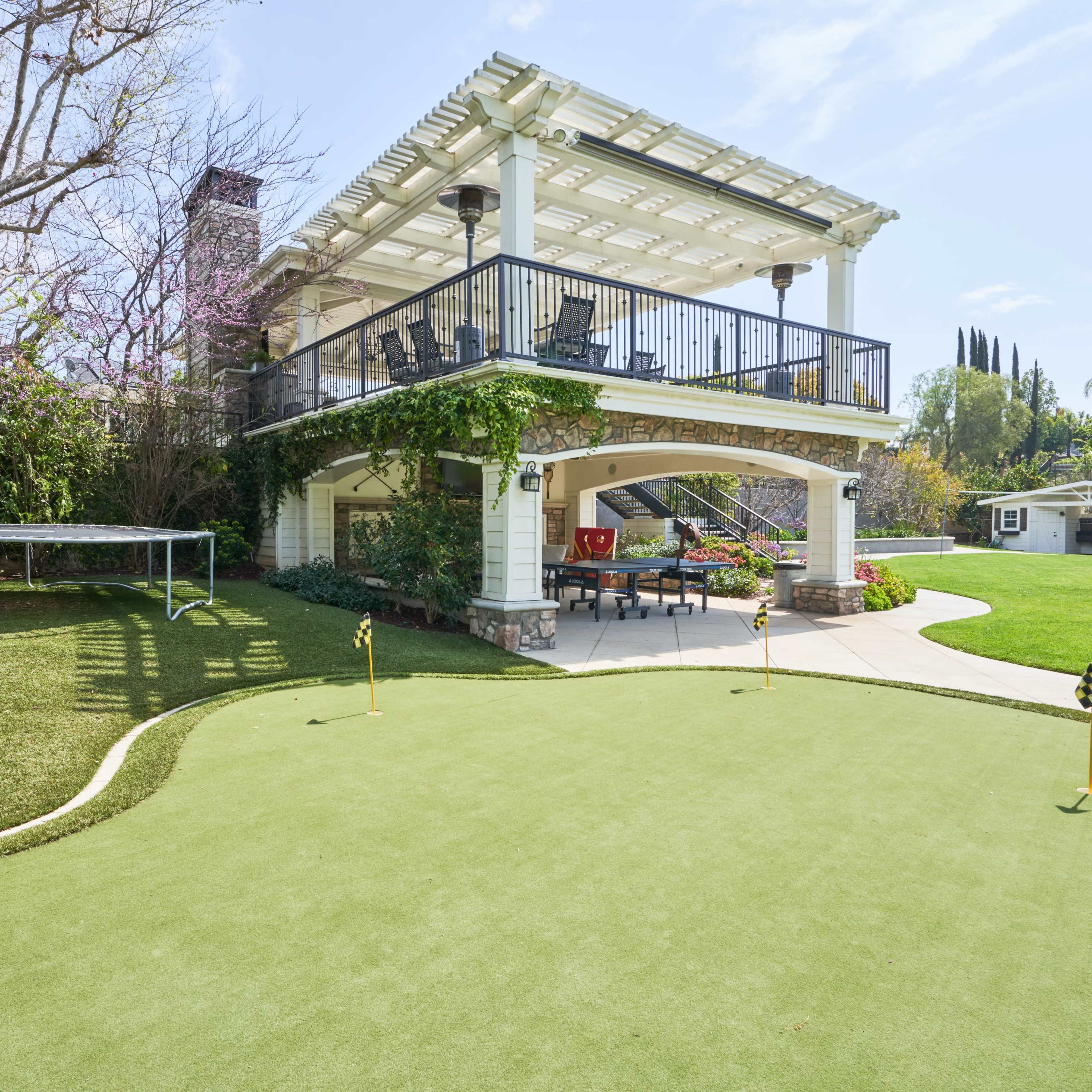 A backyard with a covered patio, a putting green, and a small soccer goal, surrounded by well-maintained landscaping.