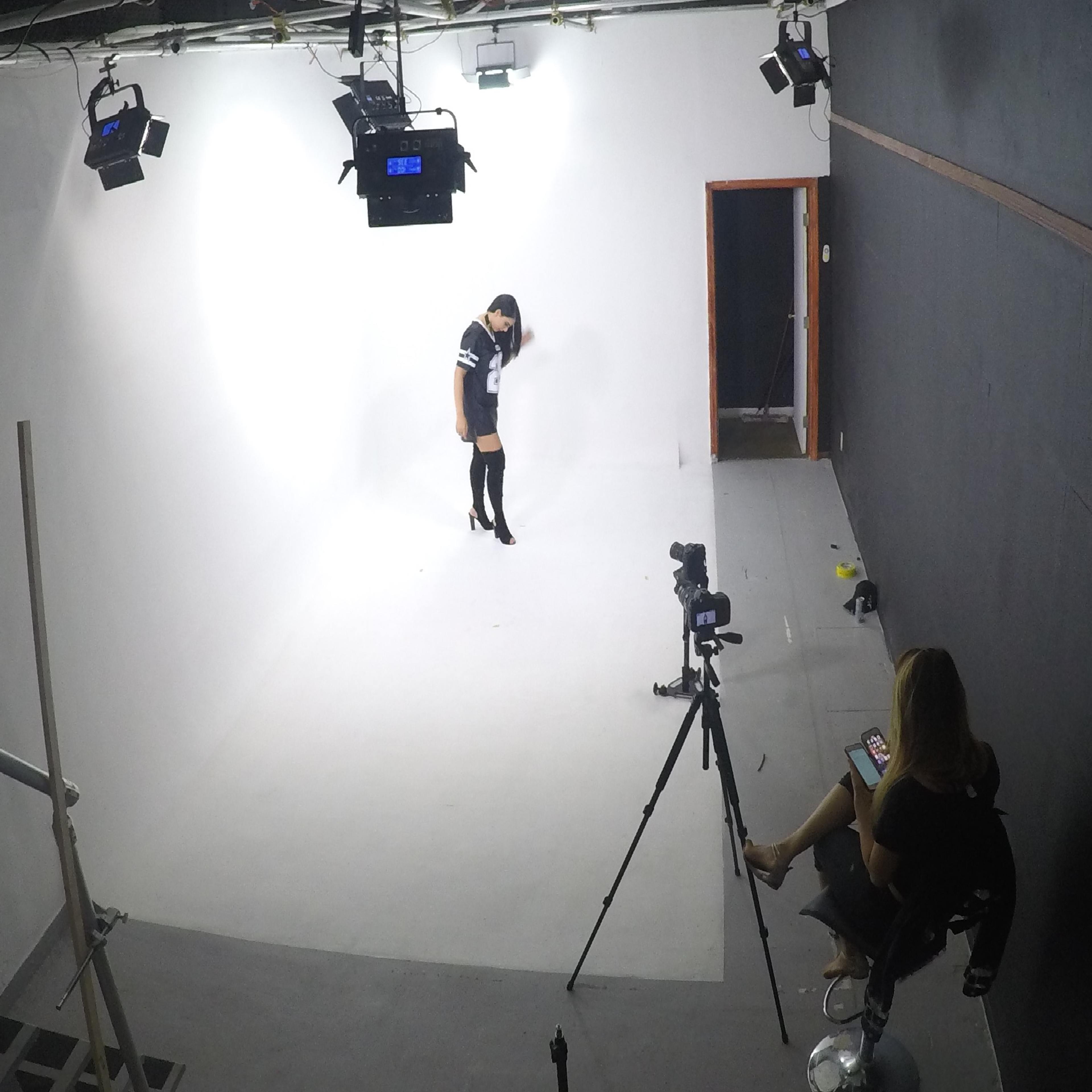 A model poses on a blank, well-lit studio set while a photographer sits nearby on a stool, using a smartphone.