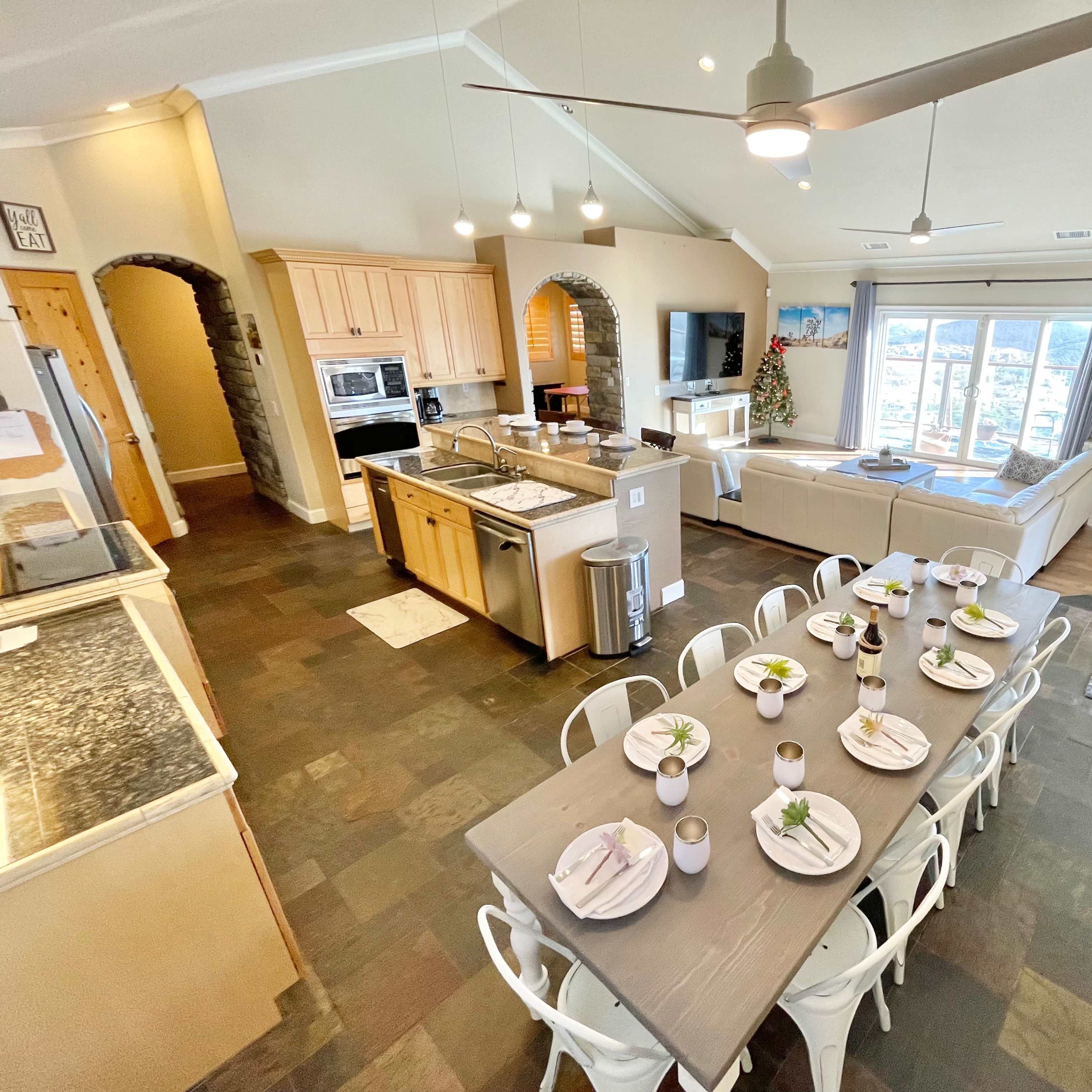 A spacious kitchen and dining area with a long table set for a meal, featuring large windows that provide views of the outdoors.