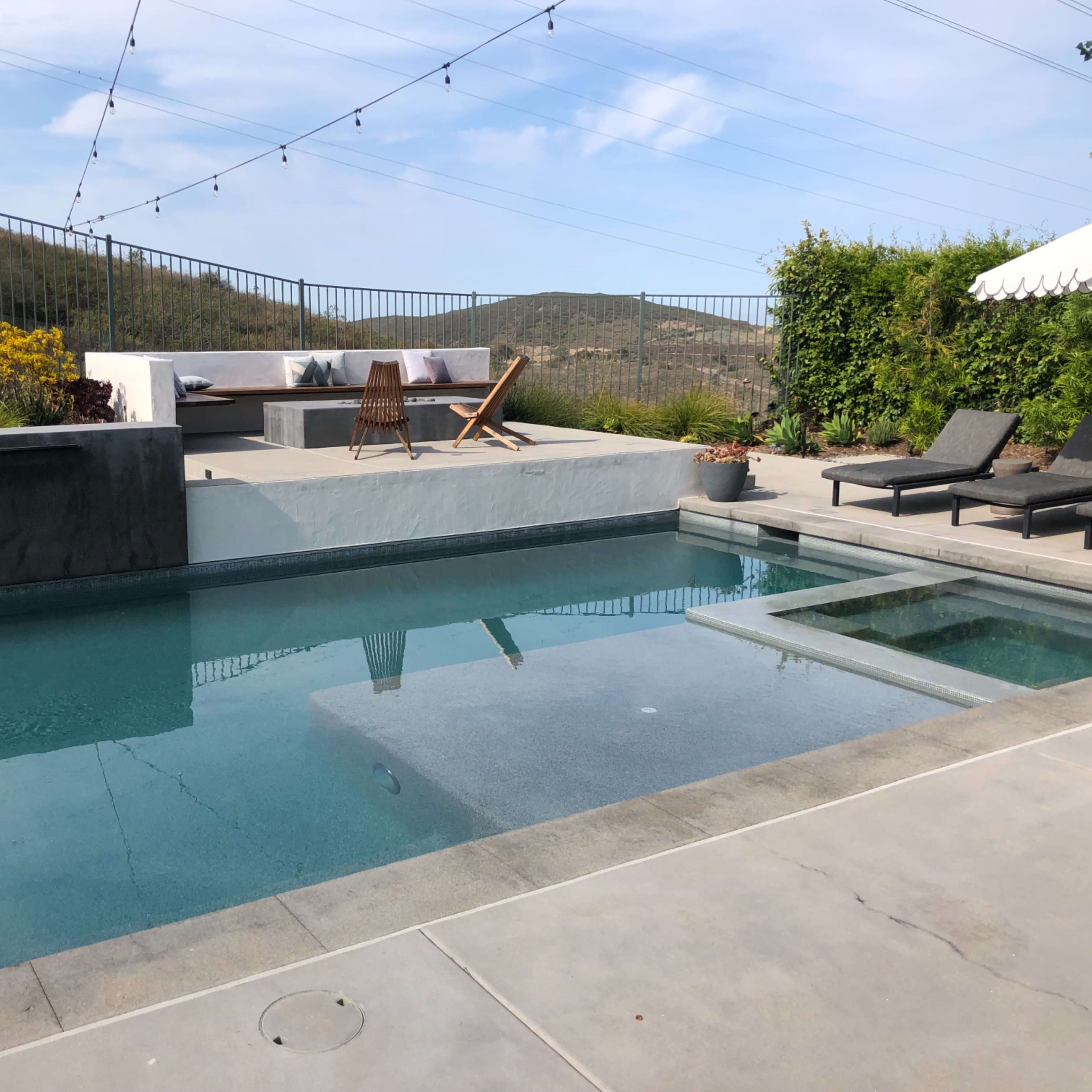 A modern backyard with a swimming pool, lounge chairs, and a seating area against a backdrop of hills and a blue sky.