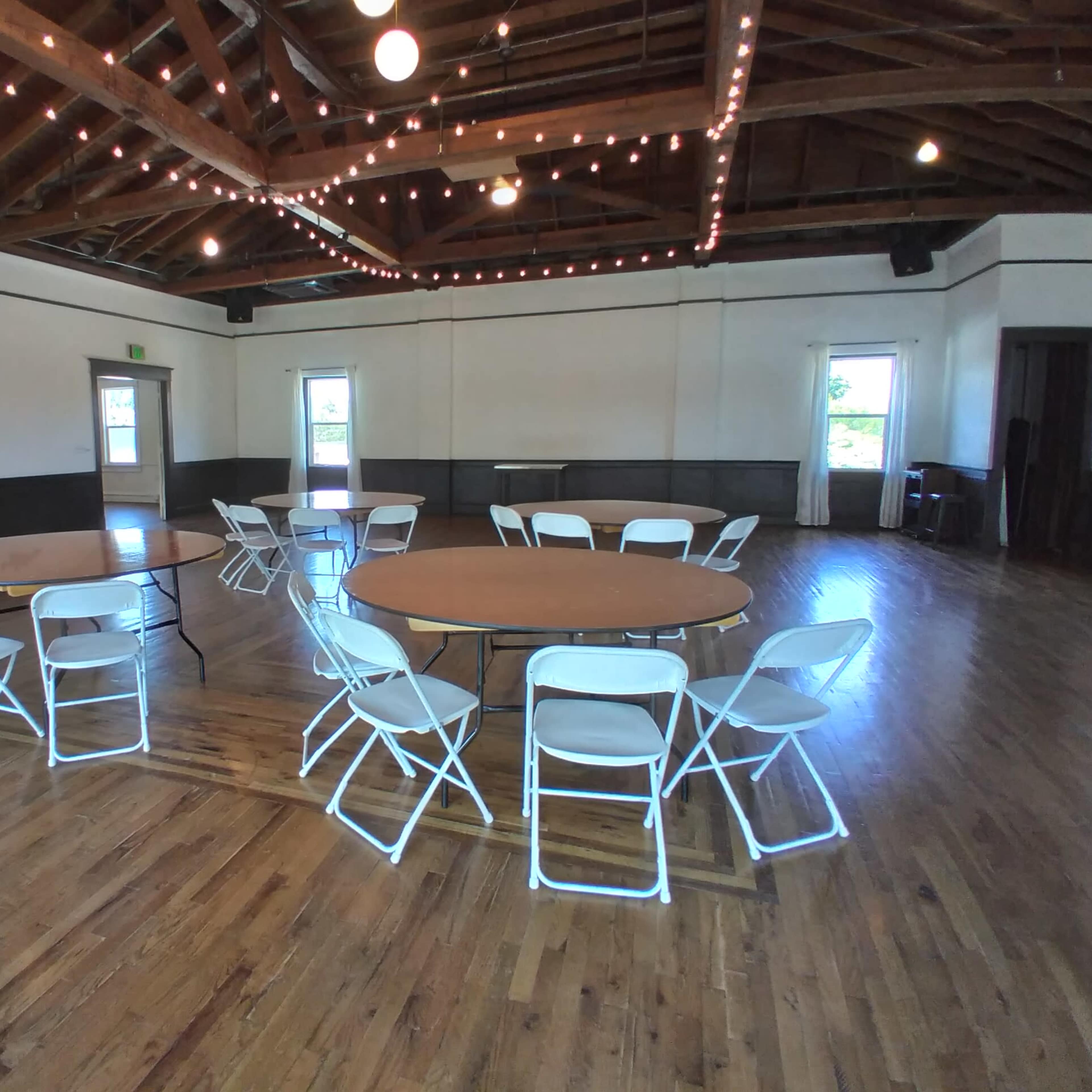 A large, well-lit room with wooden flooring, featuring several round tables surrounded by white chairs and string lights overhead.