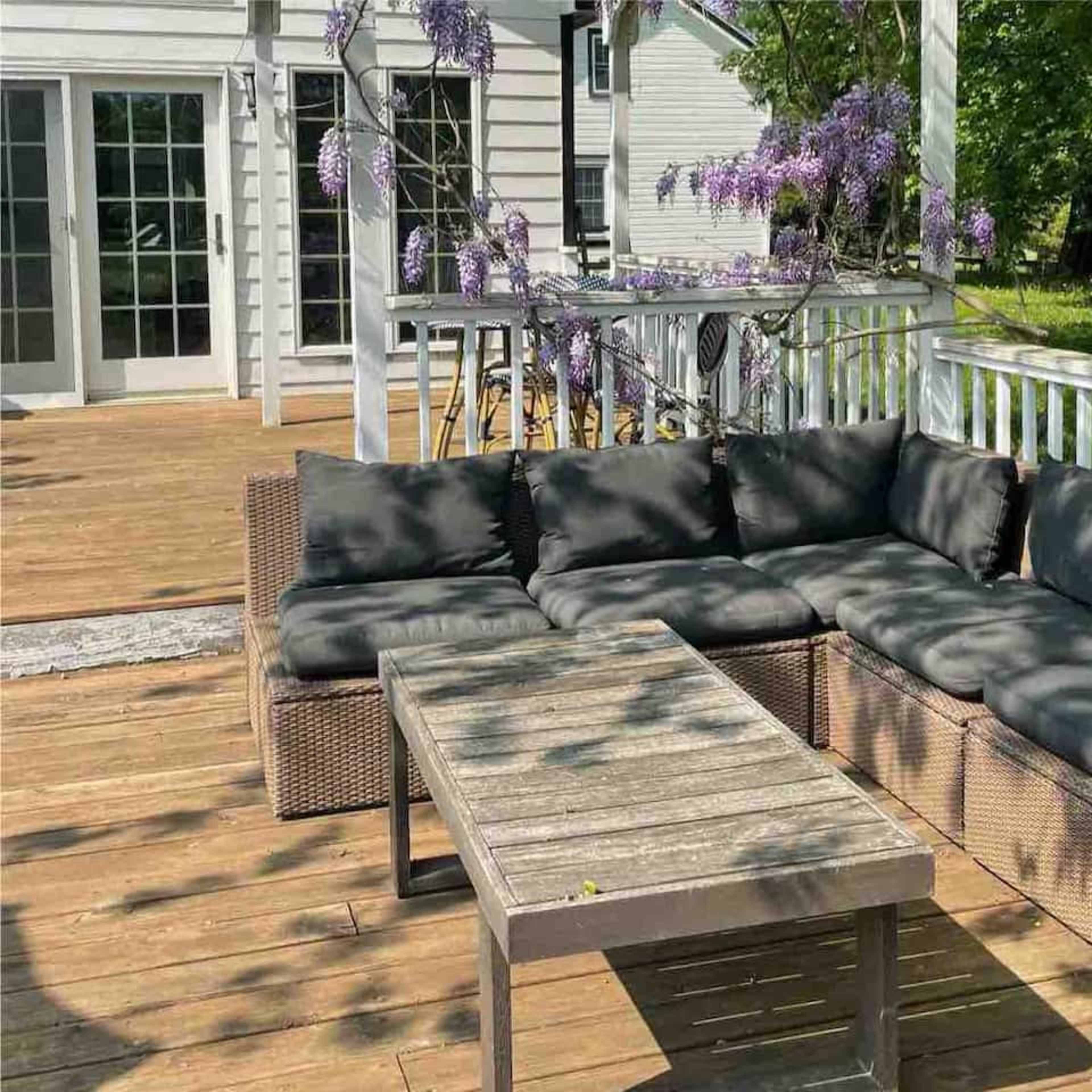 A rectangular outdoor seating area featuring a dark sectional couch and a wooden coffee table is shown on a wooden deck beneath leafy trees.