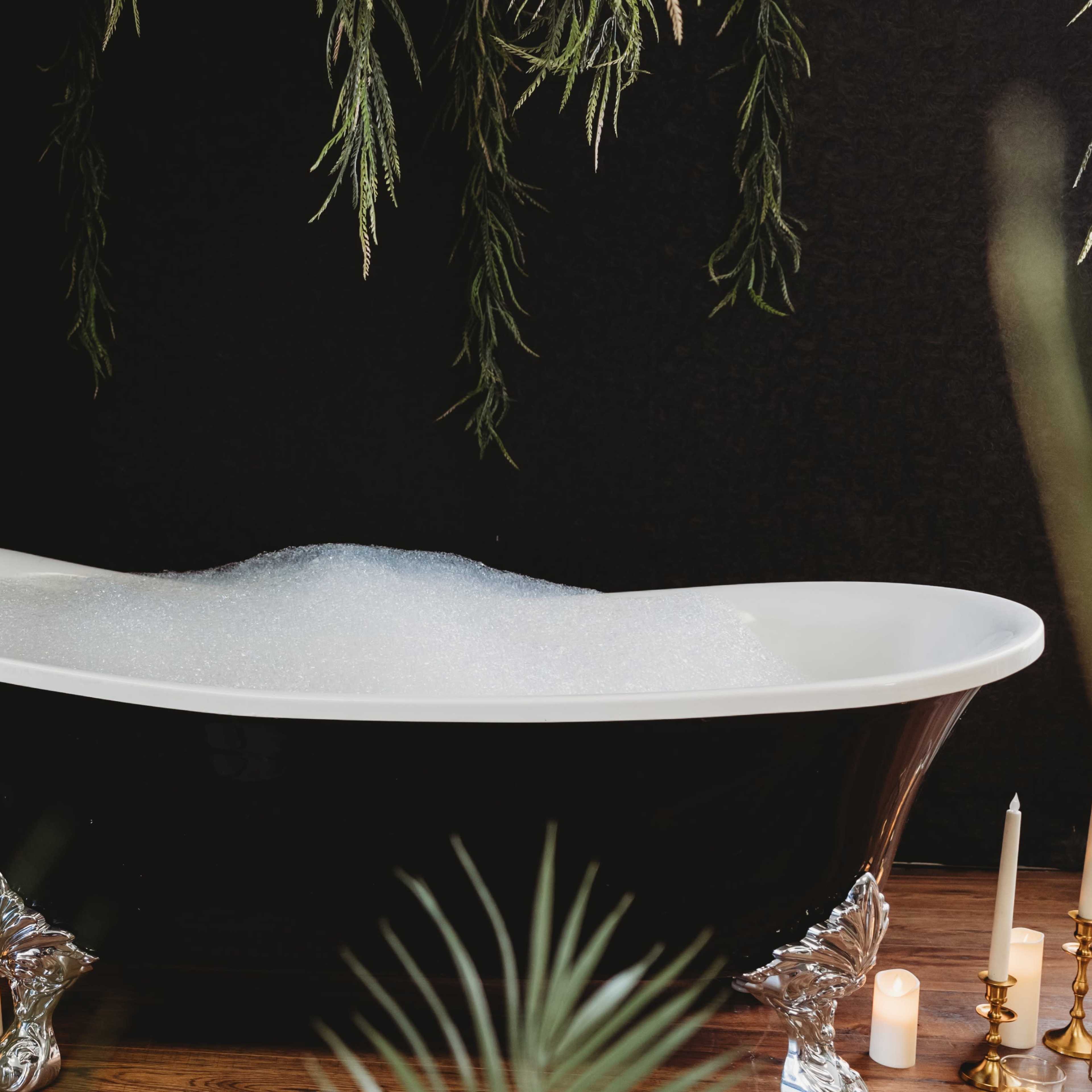 A freestanding white bathtub filled with bubbles is set against a dark background, surrounded by greenery and candles.