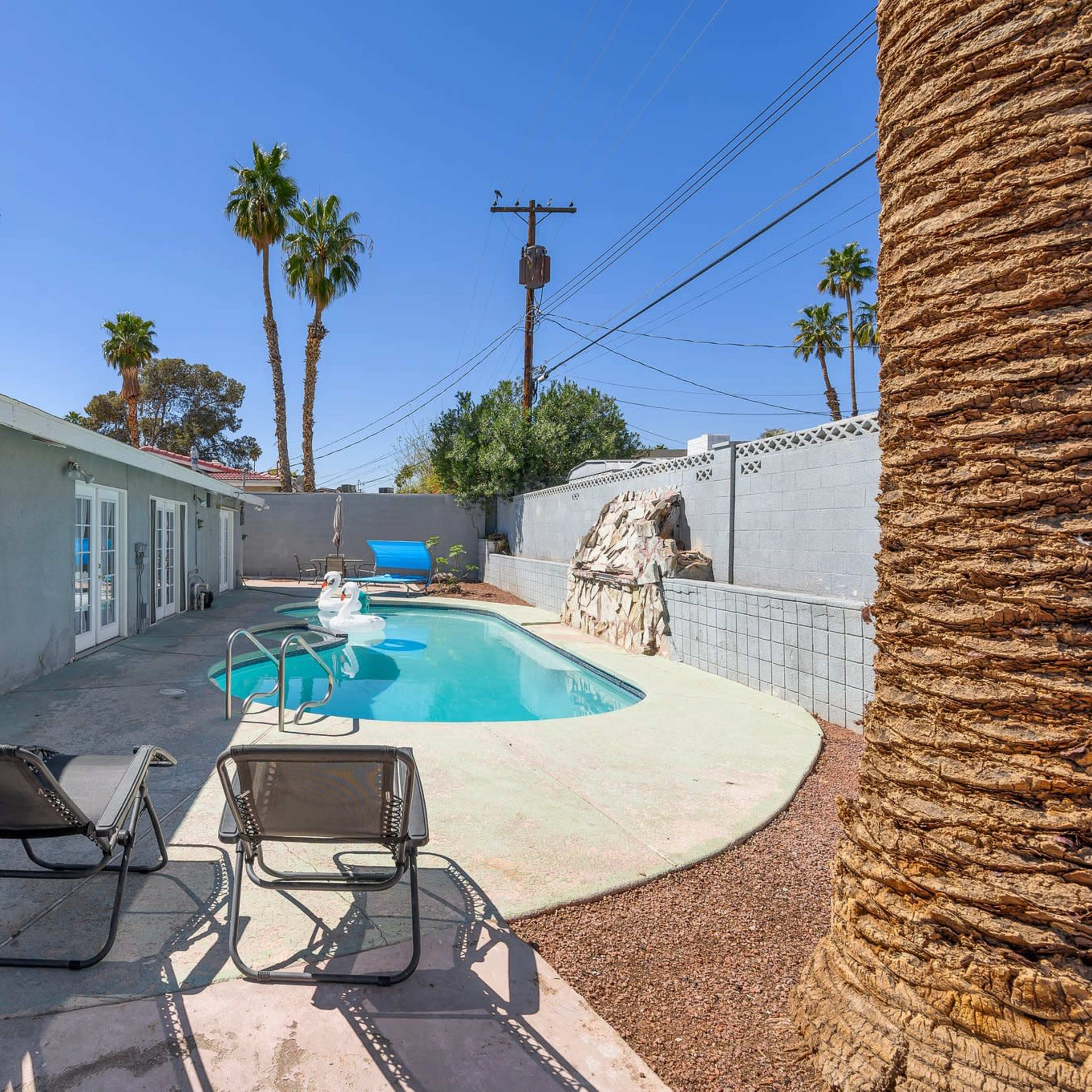 A backyard featuring a swimming pool, two lounge chairs, palm trees, and a house with a sliding glass door.