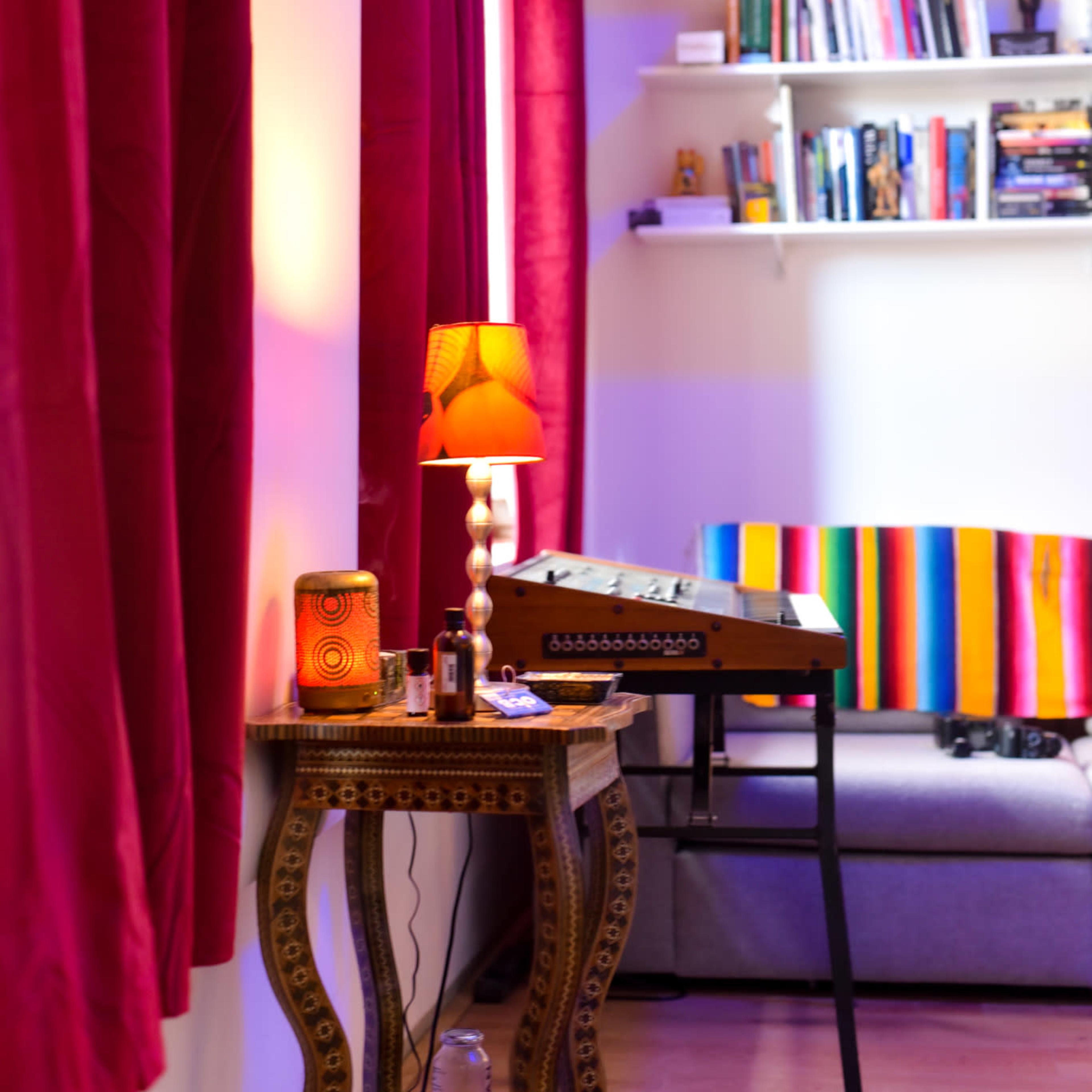 A cozy room features a decorated sofa with a colorful throw and a small table with a lamp, surrounded by red curtains and a shelf filled with books.