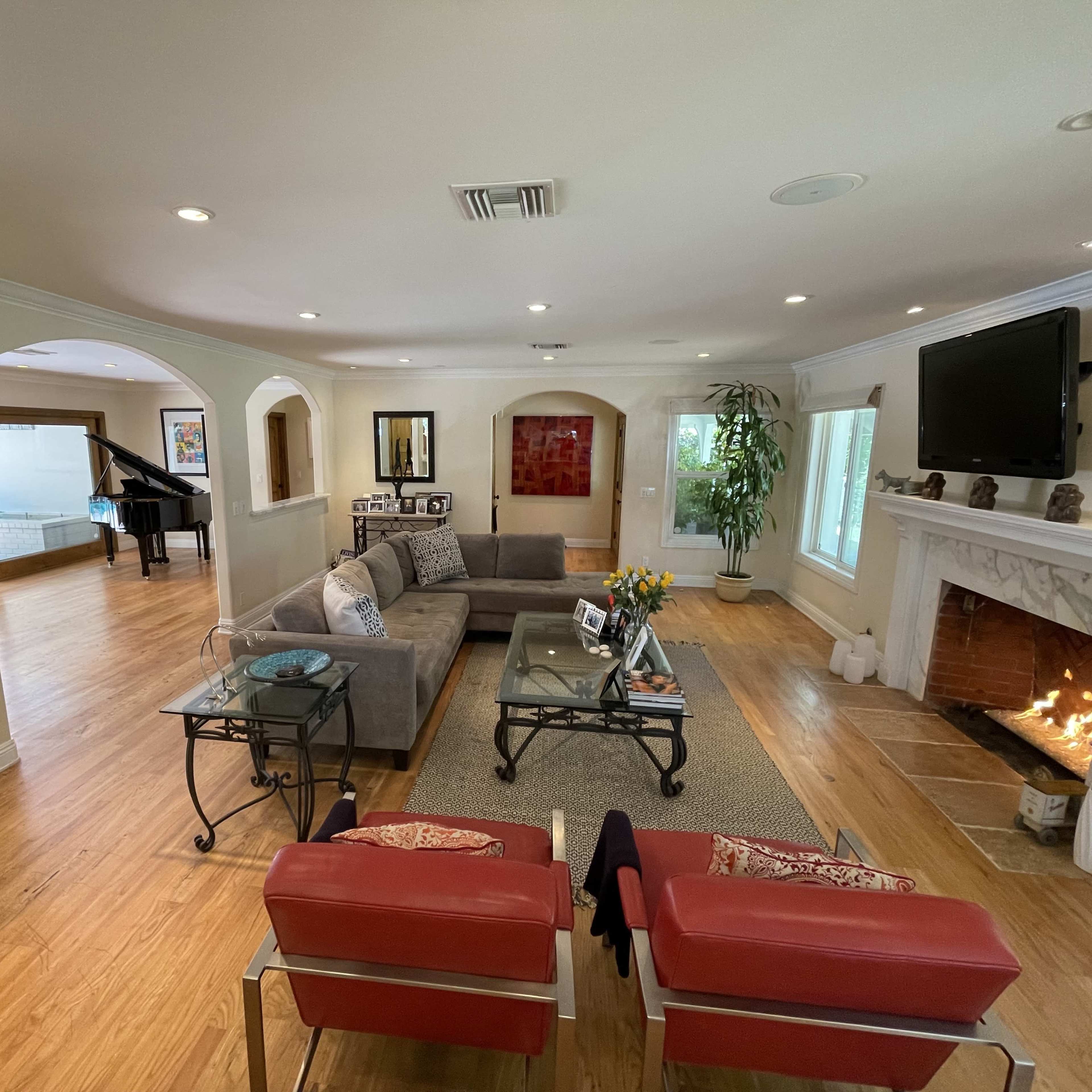 A spacious living room with a gray sectional sofa, a glass coffee table, and a fireplace, complemented by an adjacent area featuring a piano and large windows.