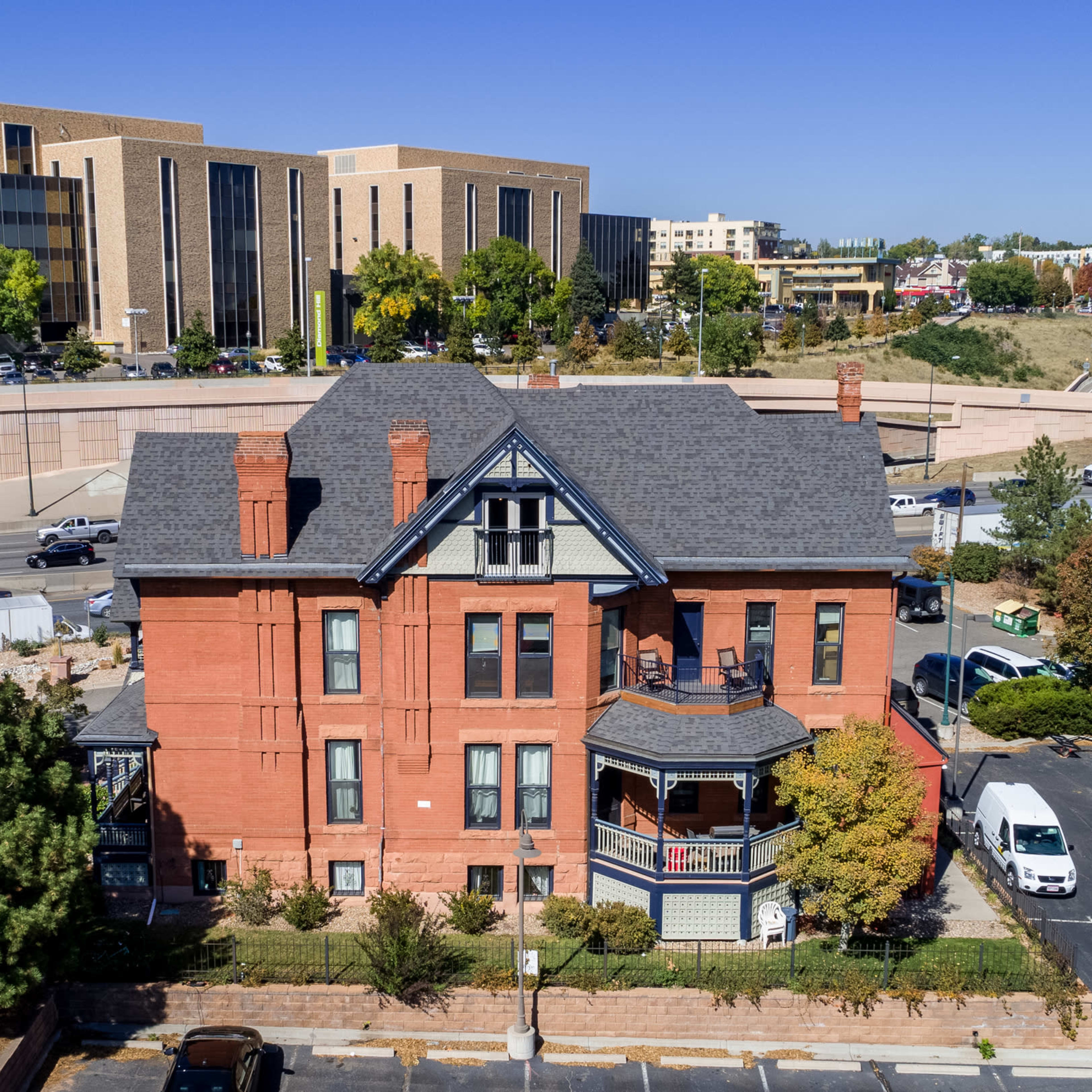 A brick mansion with a distinctive roof and balcony in the foreground, surrounded by modern buildings and a busy roadway in the background.