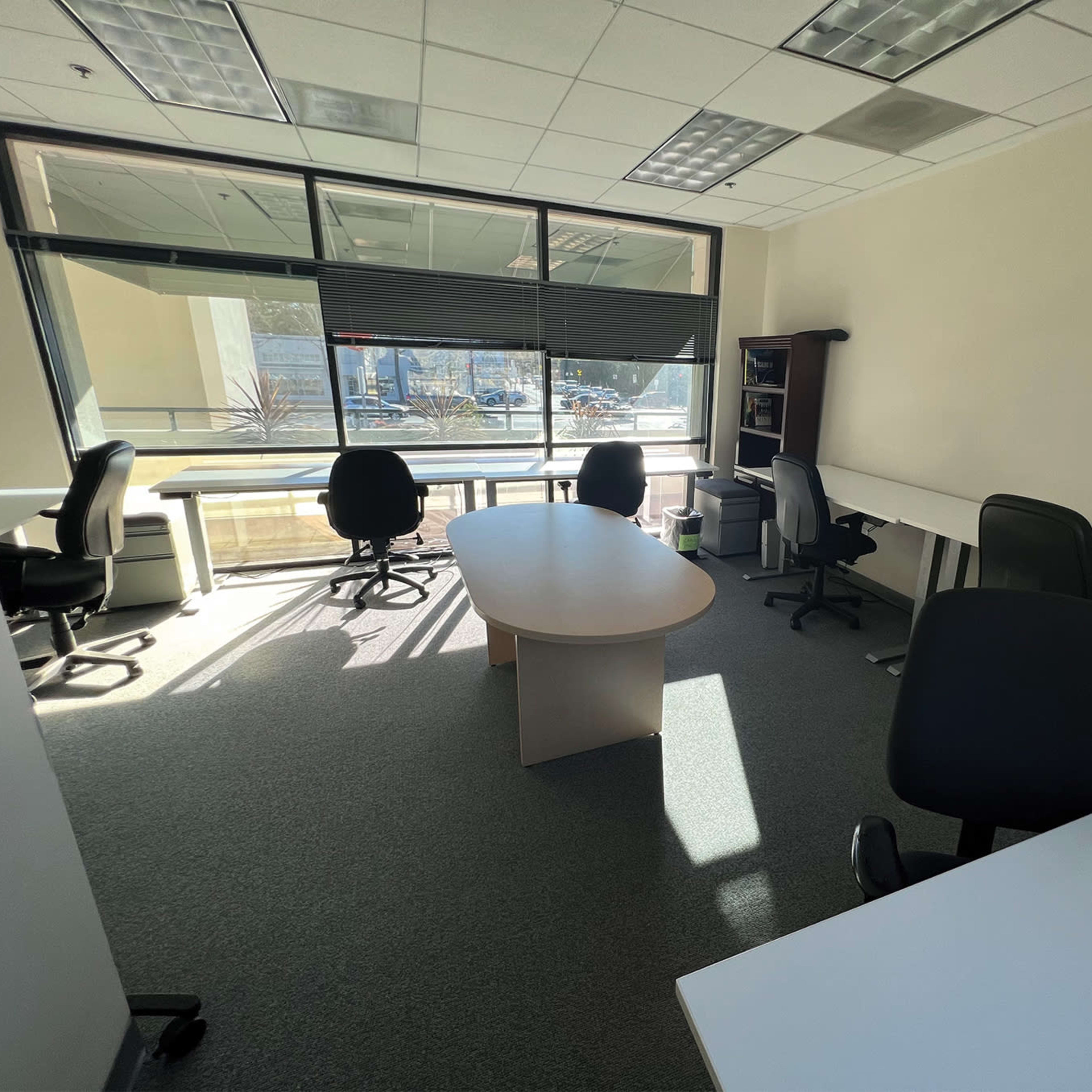 A bright office space featuring several desks and chairs, with large windows providing a view outside.