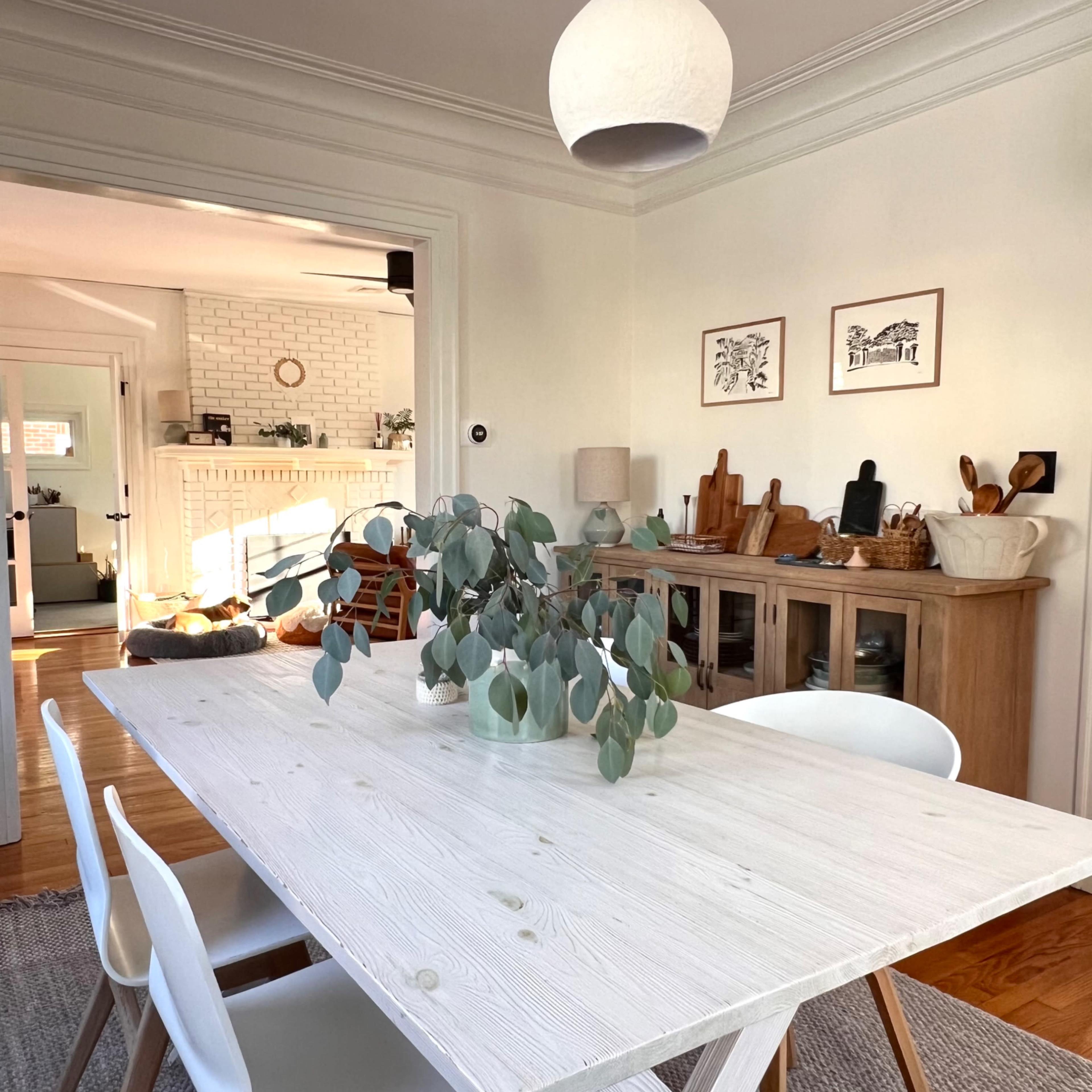 A dining table with a white surface and several chairs is positioned in a room with a light fixture and decorative elements in the background.