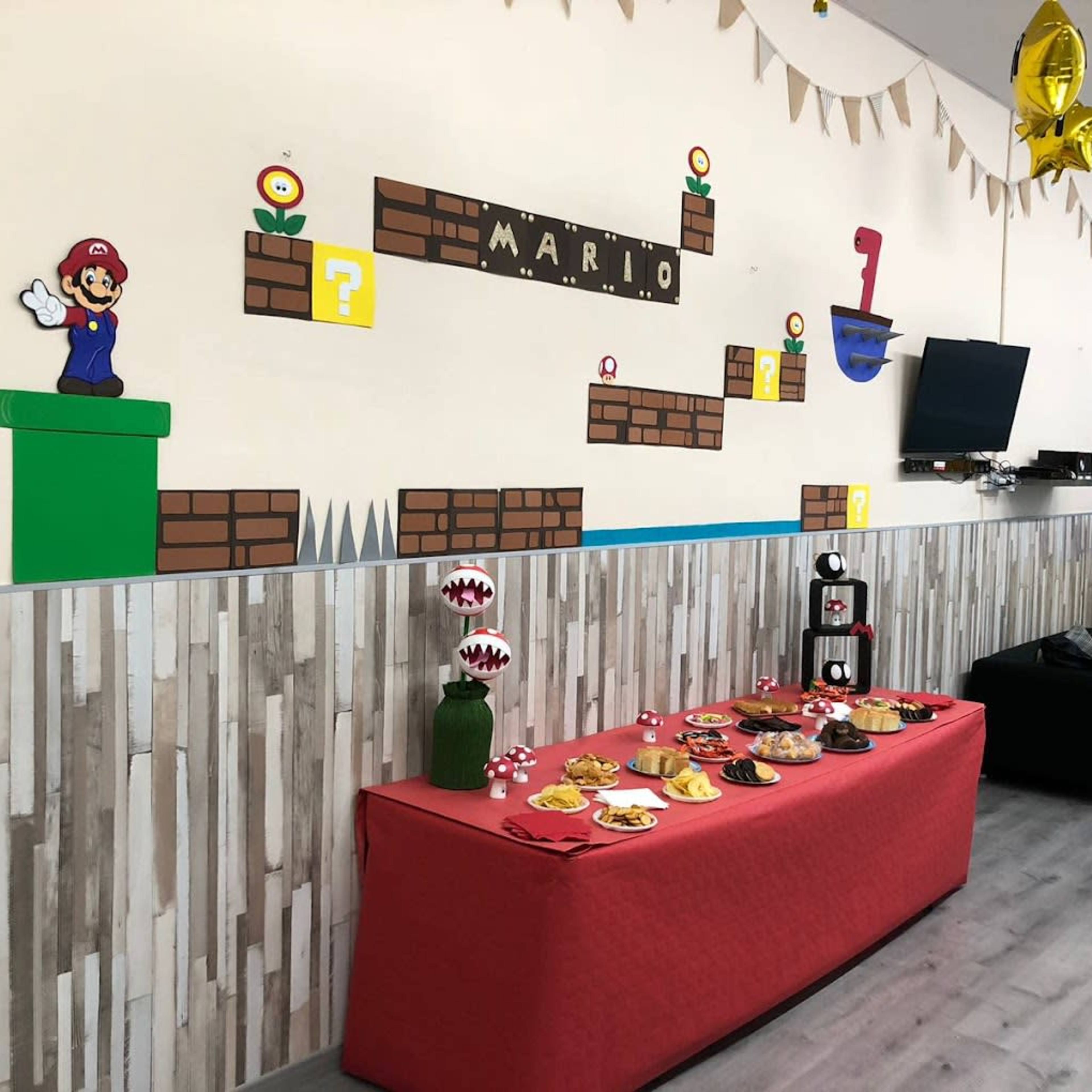 A decorated room features a Mario-themed wall display with game characters and blocks, alongside a table set with snacks and a black couch.