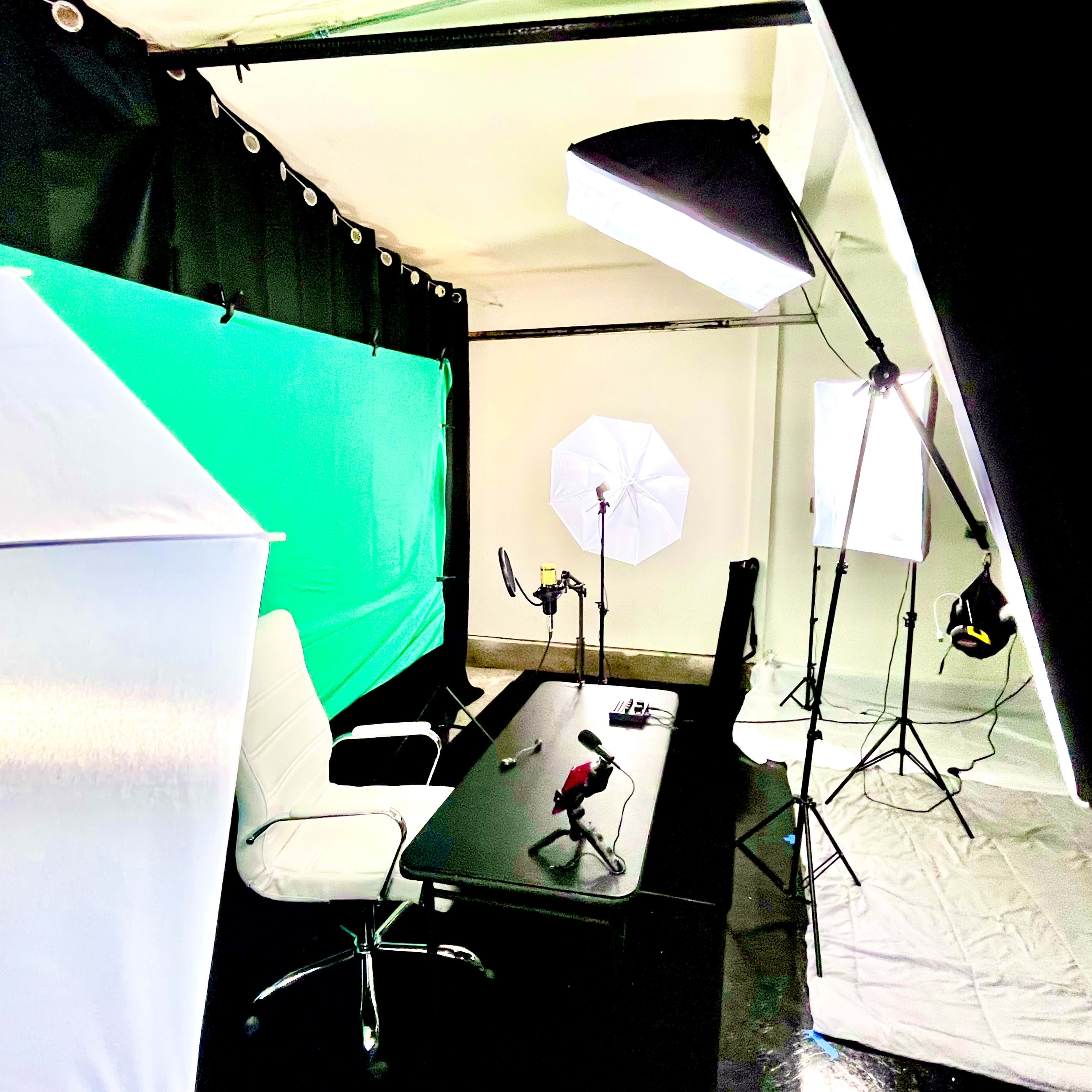 A video recording setup with a black desk, a white chair, and multiple lighting sources, including softboxes, against a green backdrop.