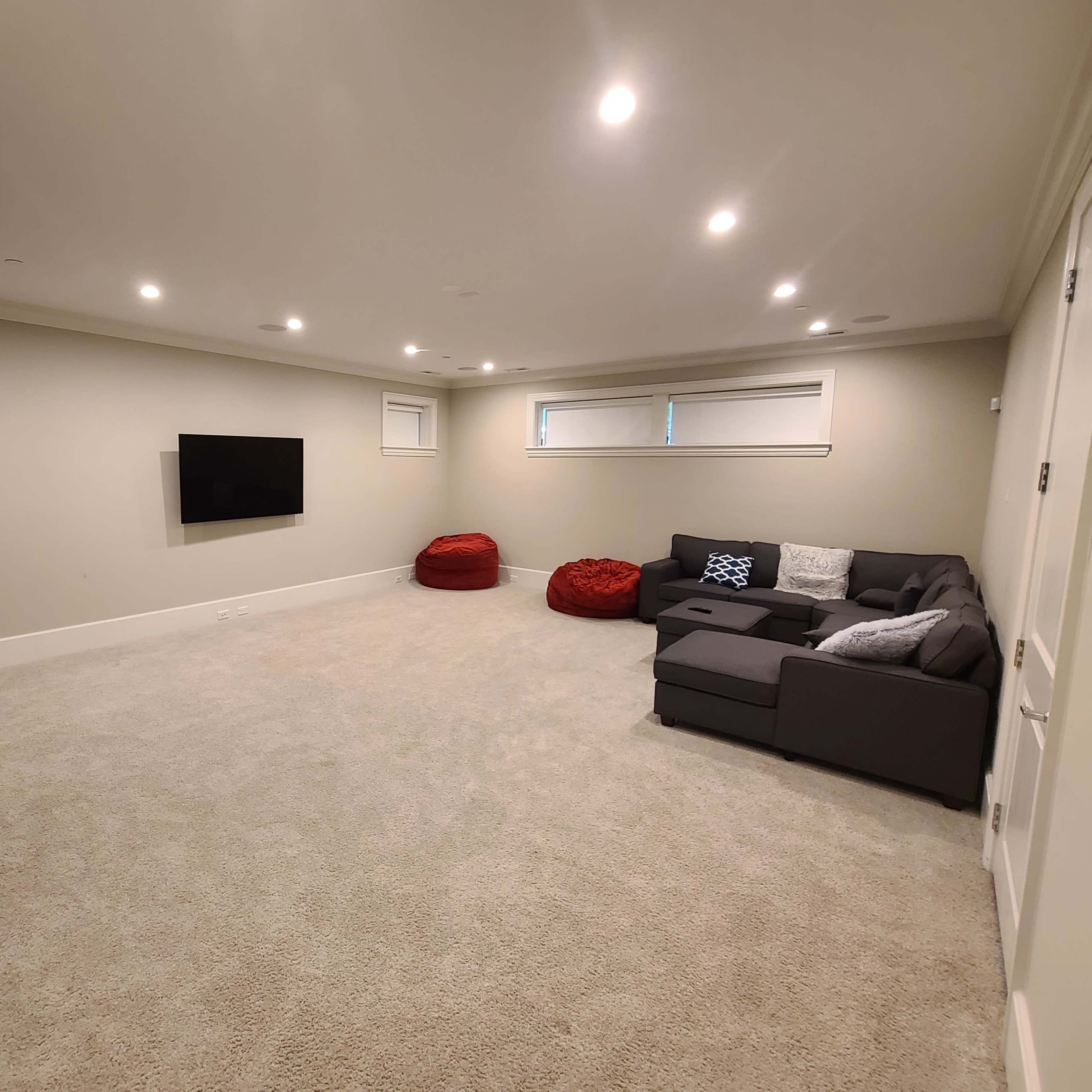 A spacious basement with beige carpet, a sectional couch, red bean bags, a mounted TV, and recessed lighting.