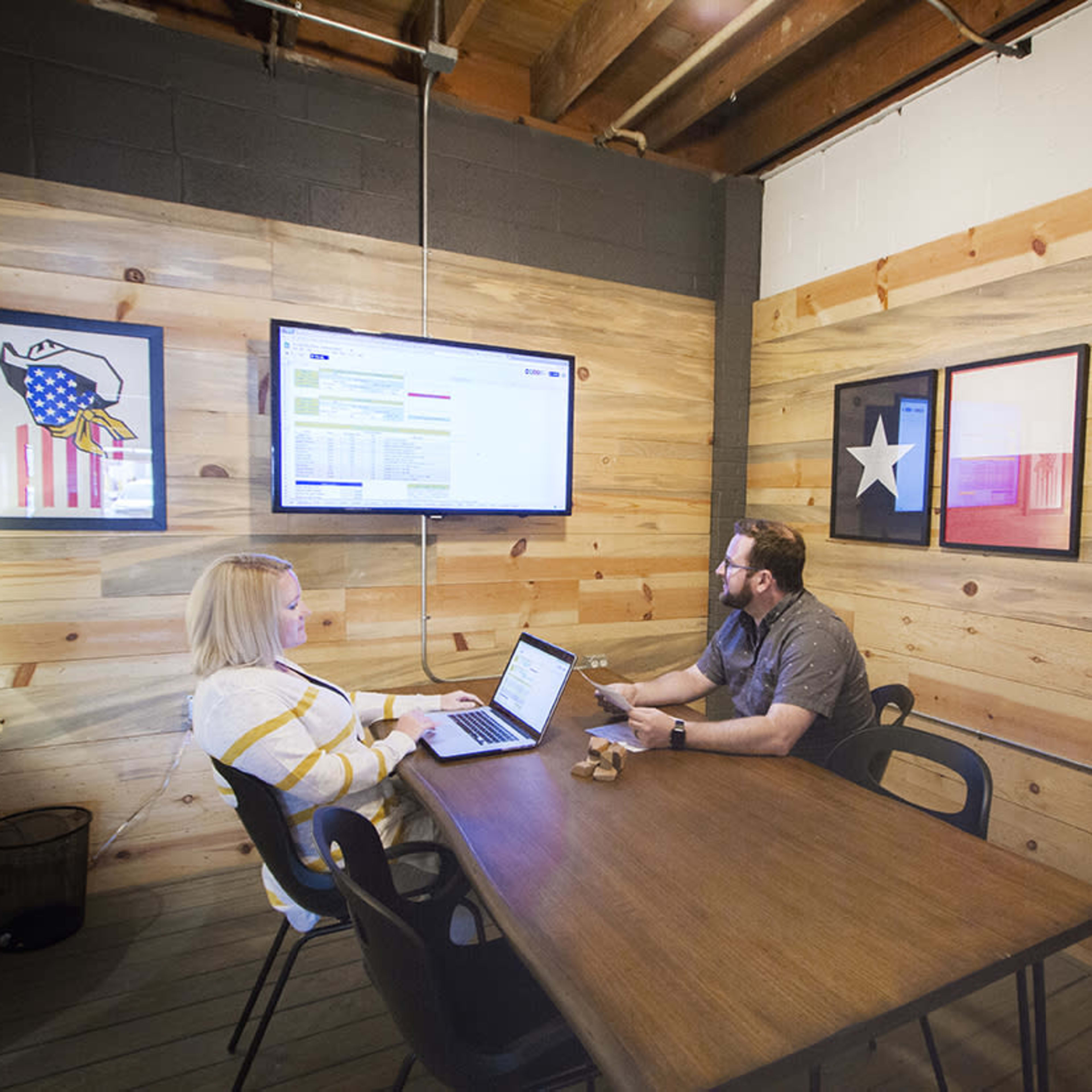 Two people are seated at a wooden table in a room with wooden walls, focusing on a laptop and a monitor displaying information.